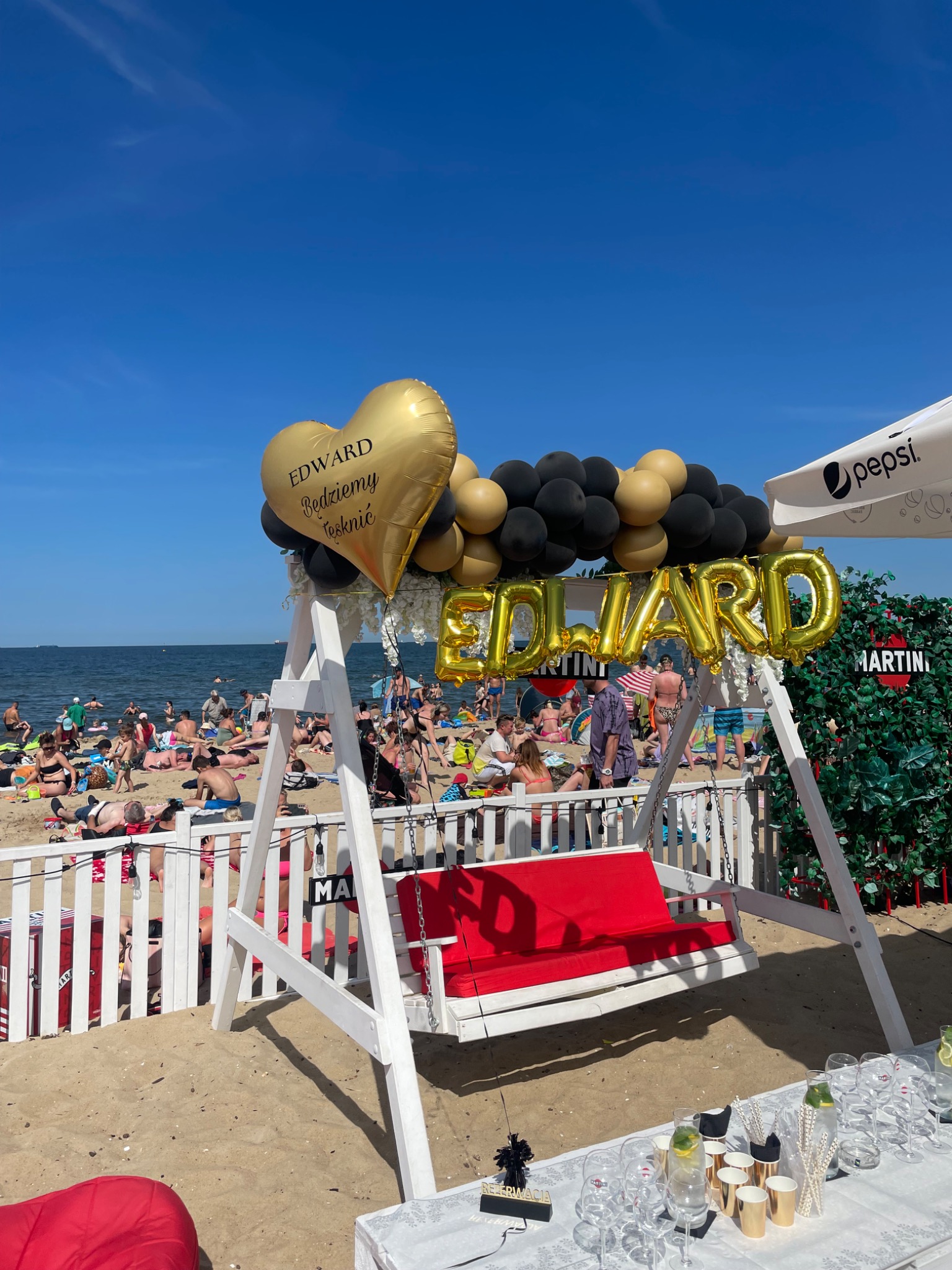 Plażowa impreza pożegnalna dla Edwarda: biała huśtawka z czerwonym siedziskiem, złote balony z napisem 'Edward Będziemy tęsknić' i 'Edward', czarno-złota girlanda balonowa, stół z napojami, tło...