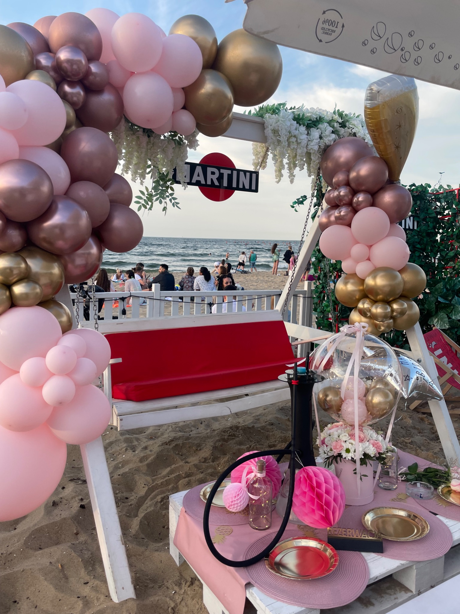 Plażowa aranżacja imprezowa w Gdańsku: huśtawka z czerwonym siedziskiem, dekoracje z różowych i złotych balonów, kwiaty, stolik z zastawą i shisha na tle morza i plażowiczów.