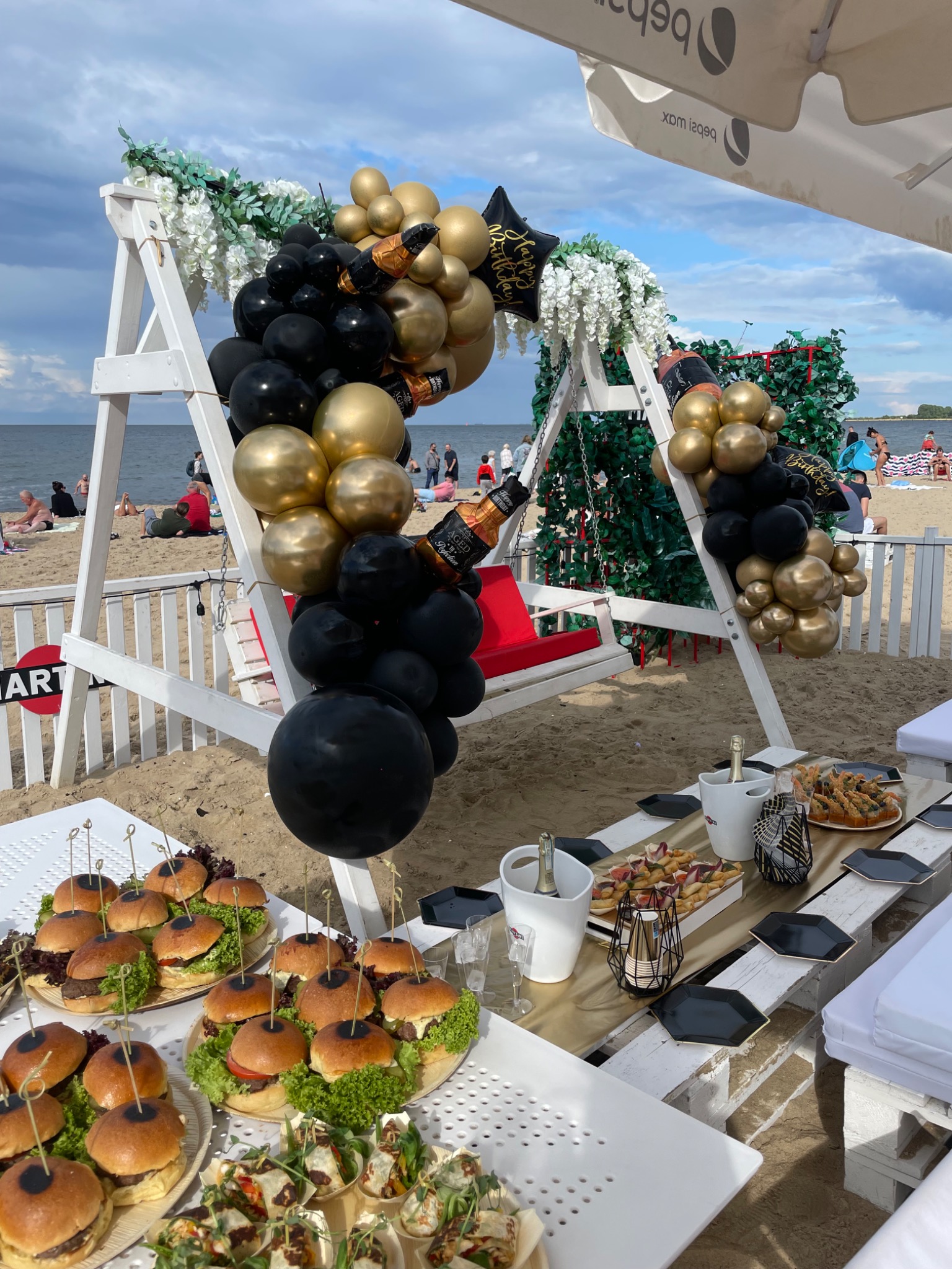 Plażowa impreza urodzinowa: biała huśtawka udekorowana czarno-złotymi balonami i kwiatami, stół z przekąskami (mini burgery, tortille) i szampanem, w tle morze i plażowicze.