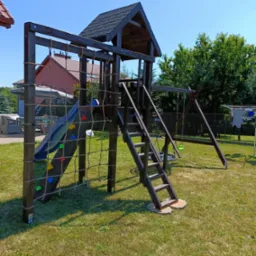 Drewniany plac zabaw z domkiem, zjeżdżalnią, ścianką wspinaczkową i huśtawką na trawniku w ogrodzie, w tle dom jednorodzinny.