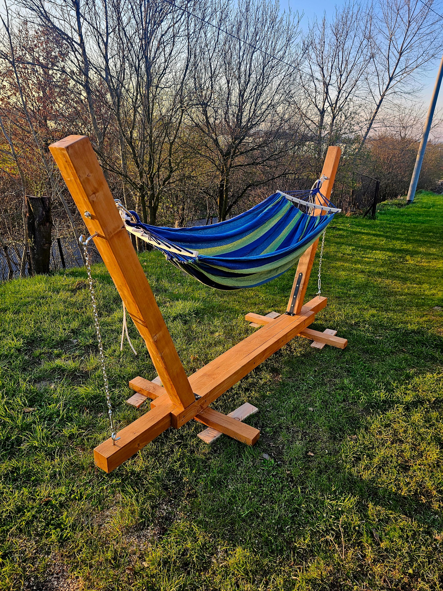 Drewniany stelaż do hamaka z niebiesko-zielonym hamakiem w paski, ustawiony na trawniku w ogrodzie, widok z bliska na konstrukcję i otoczenie.