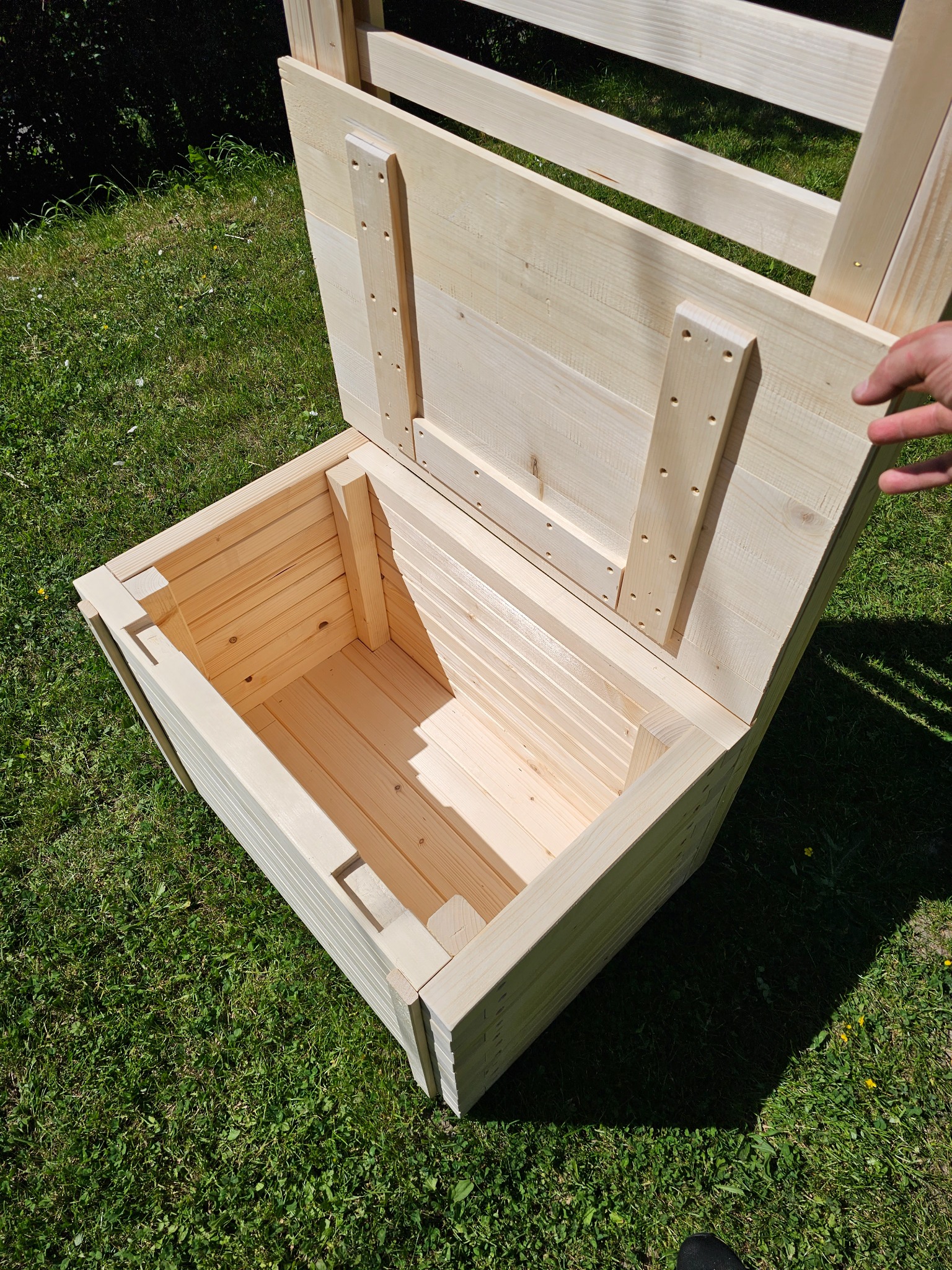 Otwarta, surowa drewniana skrzynia na trawie, widoczne wewnętrzne deski i konstrukcja wieka, dłoń przytrzymuje wieko.