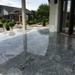 Błyszcząca, szara posadzka z płytek imitujących kamień na zewnątrz budynku, z widocznym odbiciem kolumn i nieba, otoczona zadbanym ogrodem z elementami małej architektury.