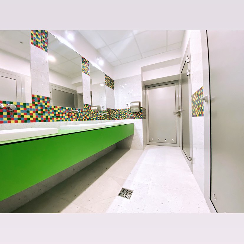 Wnętrze publicznej łazienki z długim, zielonym blatem pod umywalki, kolorową mozaiką na ścianie, dużymi lustrami i jasną, marmurową podłogą z ozdobną kratką odpływową.