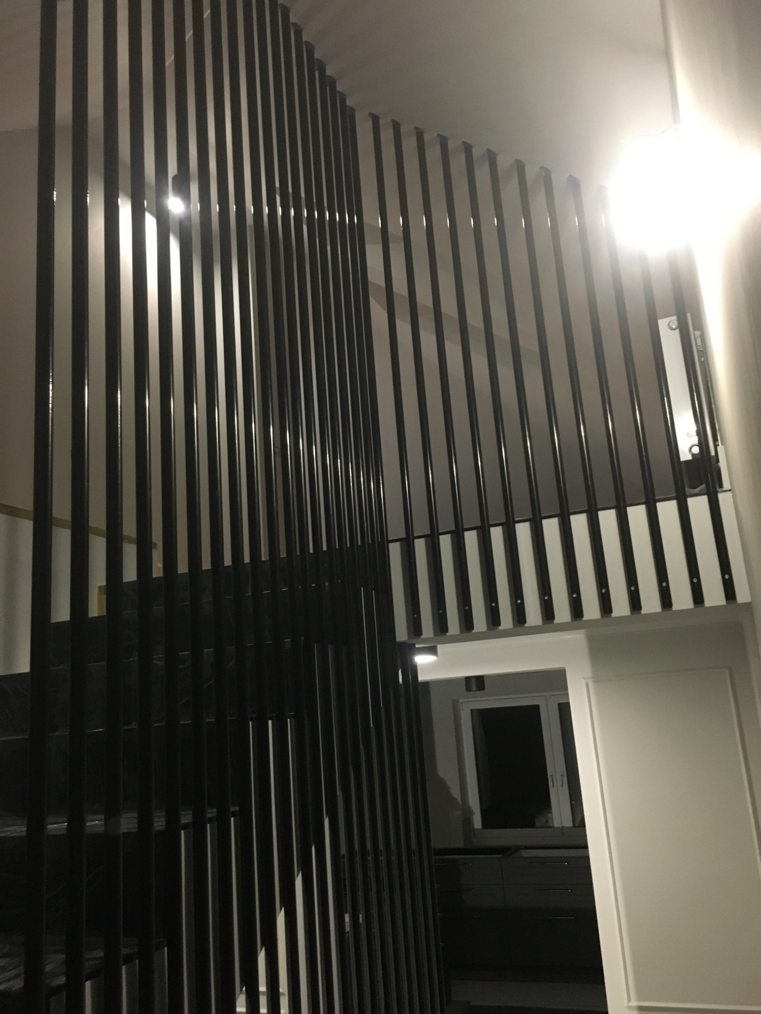 Nowoczesne schody z czarnymi, pionowymi balustradami, widok z dołu, białe ściany z dekoracyjnymi listwami, widoczny fragment kuchni w tle.