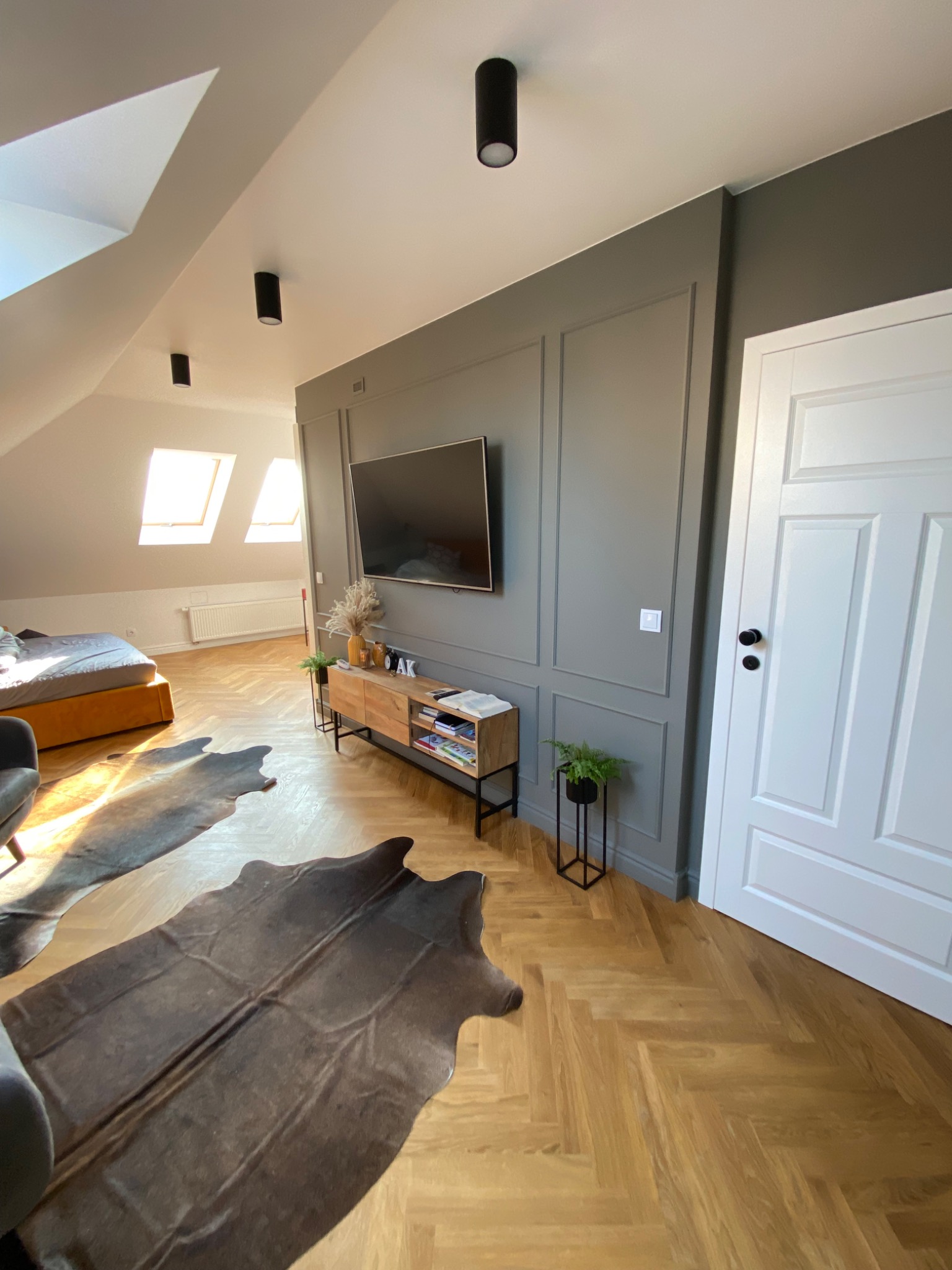 Pokój na poddaszu z drewnianą podłogą w jodełkę, telewizorem na ścianie z szarą boazerią, drewnianą komodą z dekoracjami i dywanami imitującymi skórę zwierzęcą.