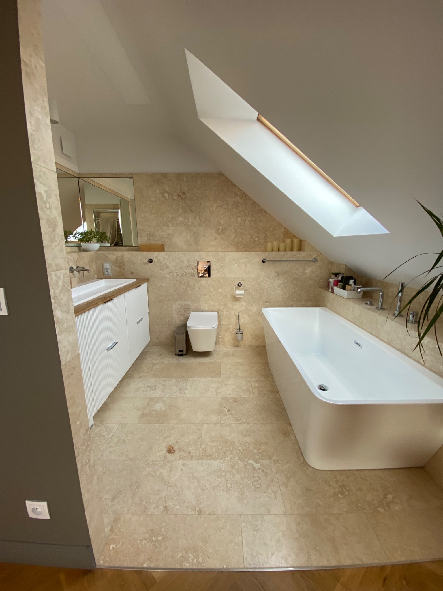 Nowoczesna łazienka na poddaszu z beżowymi płytkami, białą wanną wolnostojącą i umywalką z szafką z lamelami, oświetlona oknem dachowym.