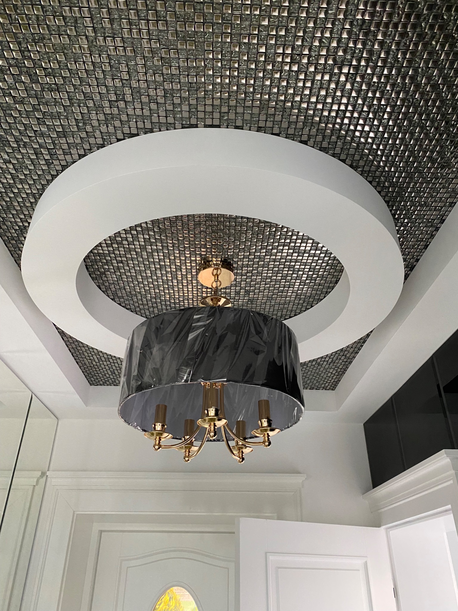 Srebrna mozaika na suficie z okrągłym białym elementem dekoracyjnym i złotym żyrandolem w czarnym abażurze, częściowo owiniętym folią ochronną.