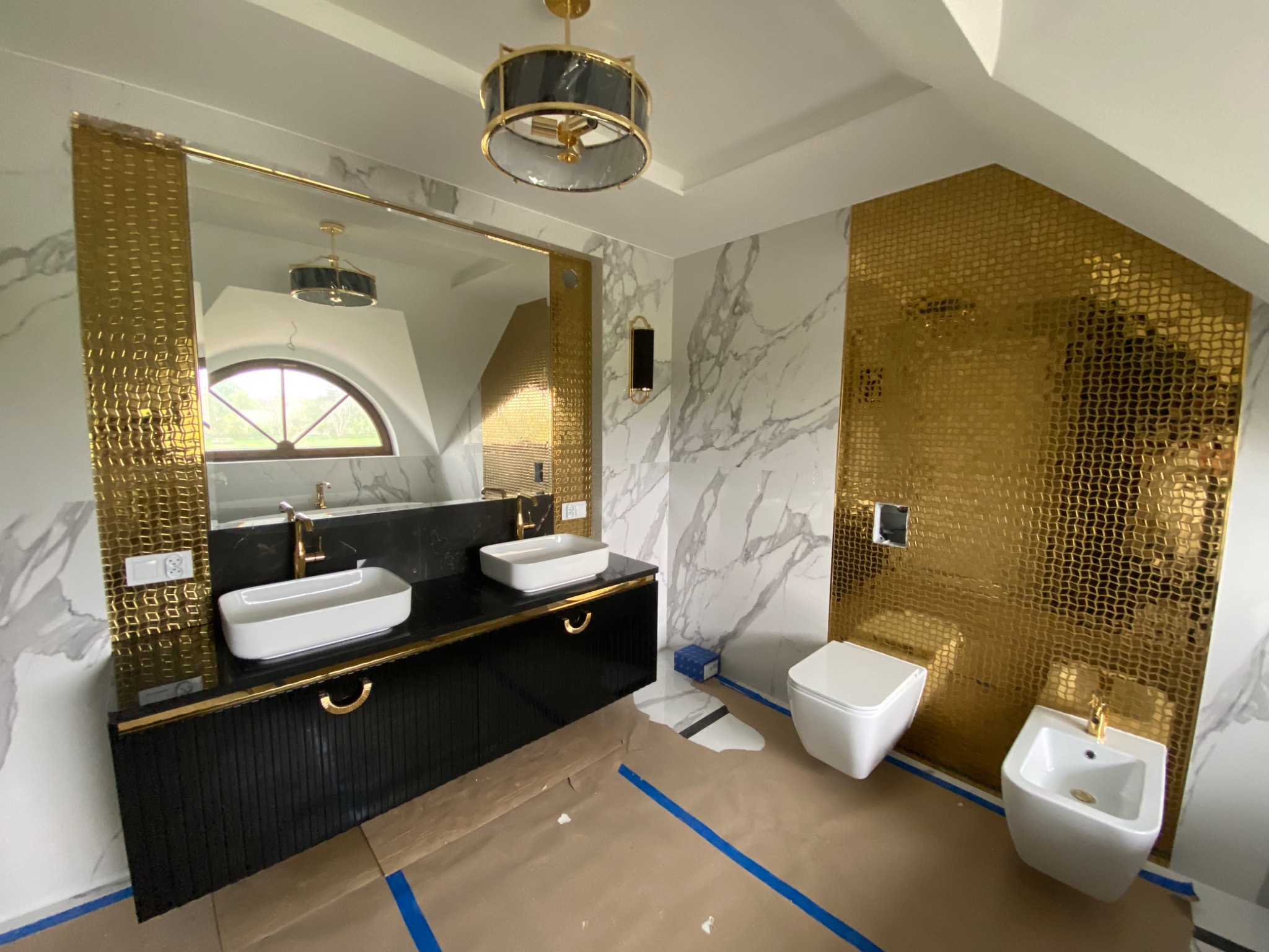 Luksusowa łazienka w Zambrowie w trakcie wykańczania: złota mozaika na ścianie, marmurowe płytki, czarna szafka z umywalkami i lustro. Podłoga zabezpieczona folią malarską.