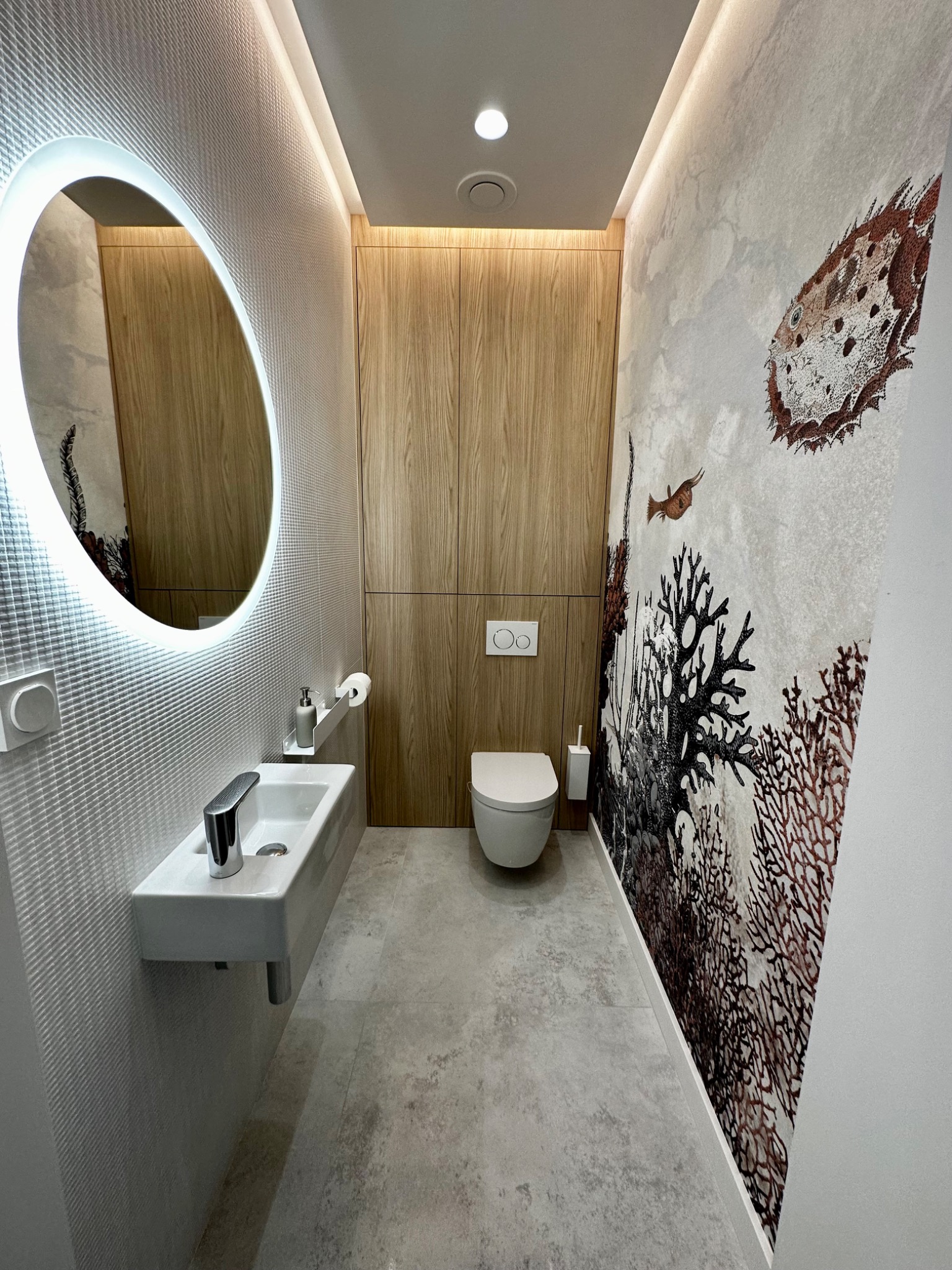 Nowoczesna toaleta z podświetlanym okrągłym lustrem, wiszącą umywalką i toaletą, drewnianą zabudową oraz tapetą z motywem morskim.