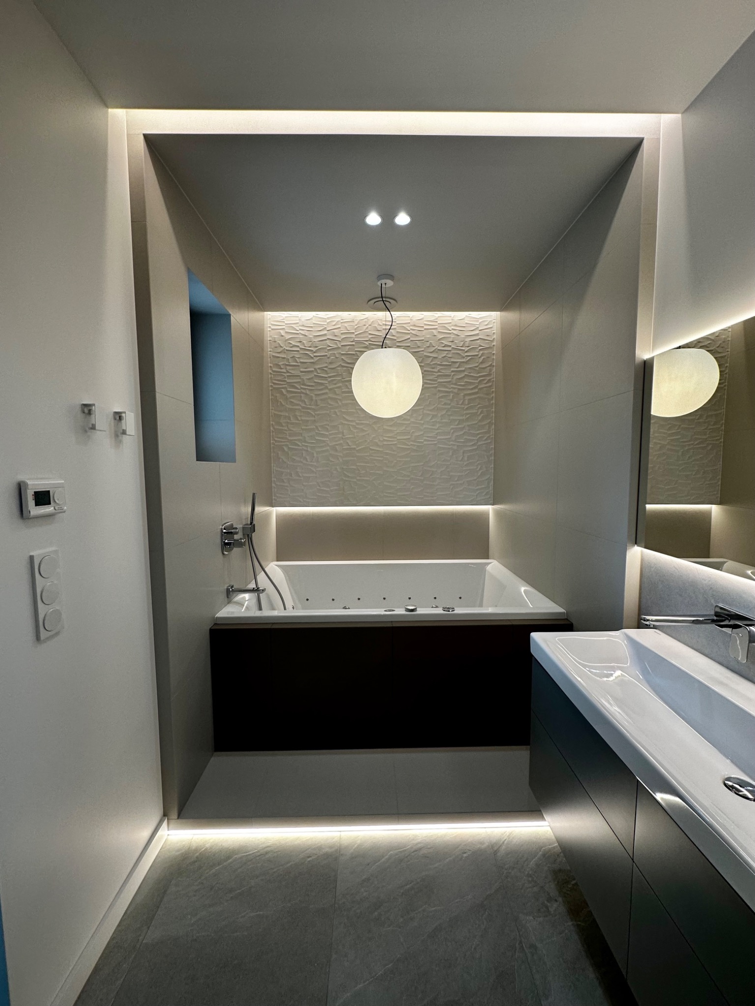Nowoczesna łazienka z podświetlaną wanną z hydromasażem, umywalką na szafce i dekoracyjną ścianą z fakturą, oświetlona lampą kulą i listwami LED.