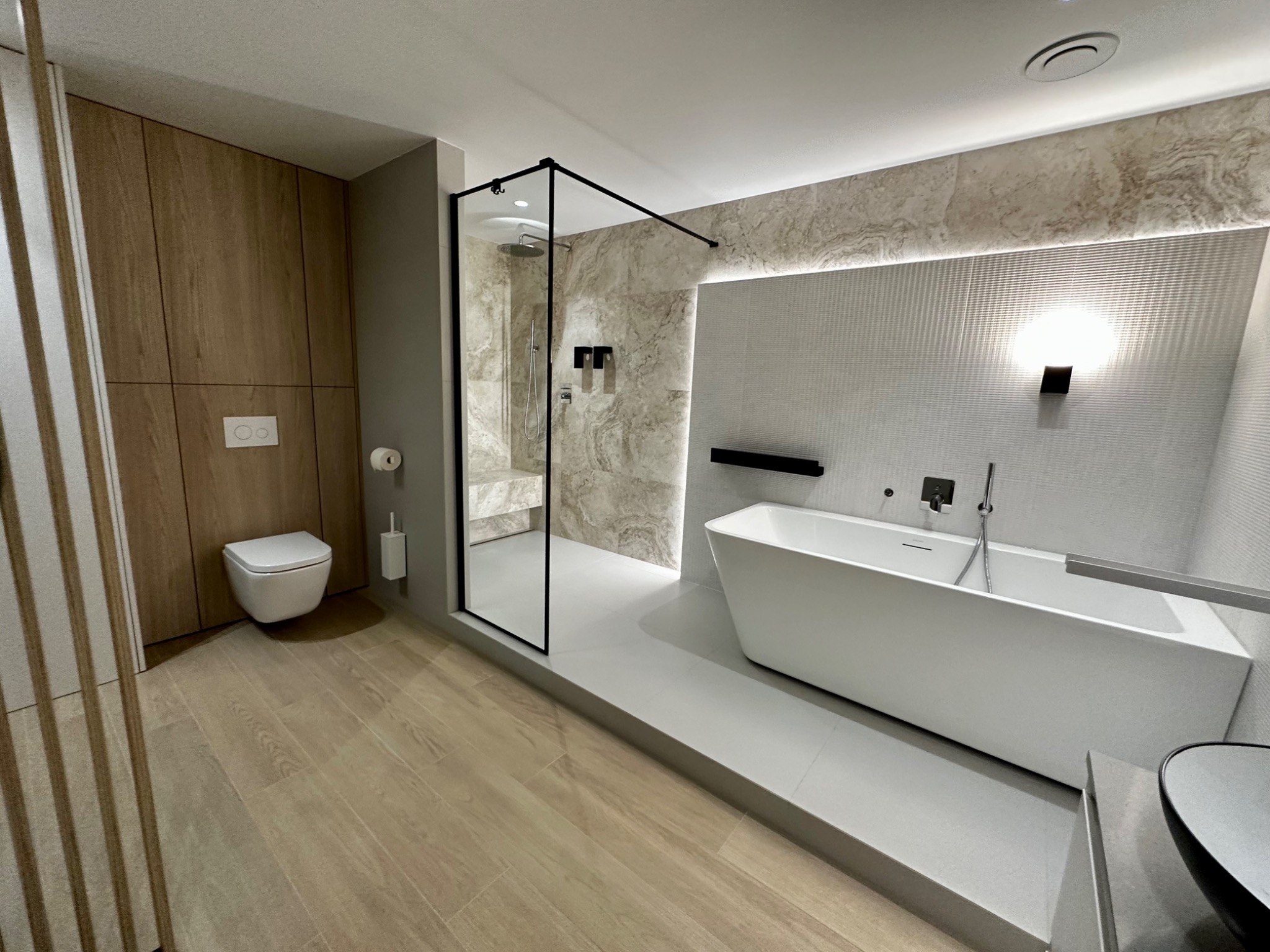 Nowoczesna łazienka z kabiną prysznicową typu walk-in, wolnostojącą wanną, podwieszaną toaletą i drewnopodobną podłogą, oświetlona lampą ścienną i listwą LED.