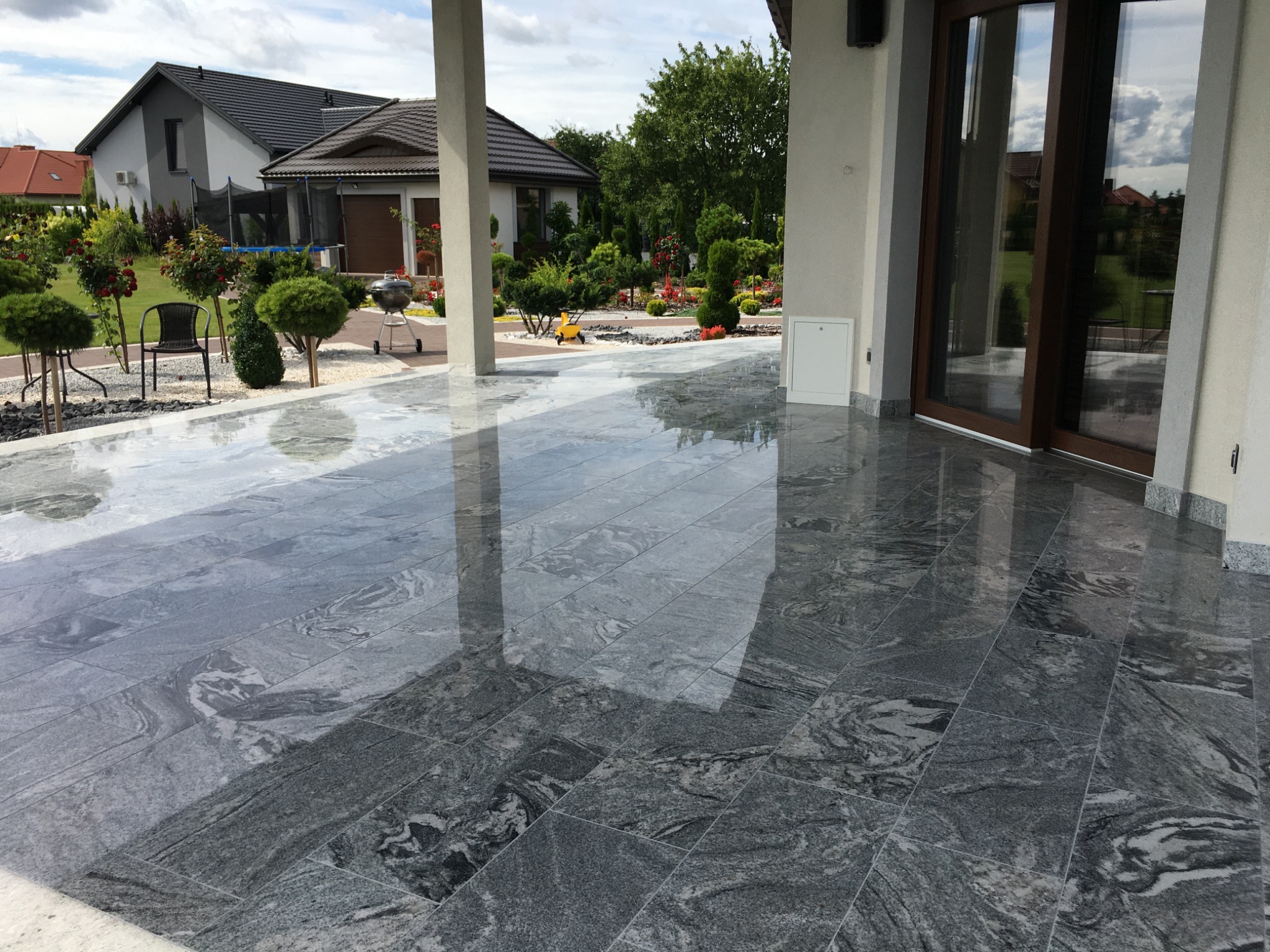 Błyszcząca, szara posadzka z płytek imitujących kamień na zewnątrz budynku, z widocznym odbiciem kolumn i nieba, otoczona zadbanym ogrodem z elementami małej architektury.