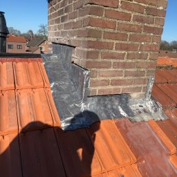 AS DAKWERKEN - Ceglany komin z obróbką blacharską na dachu z czerwonej dachówki, widok z góry w słoneczny dzień. Detal zabezpieczenia komina przed przeciekaniem.