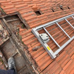 AS DAKWERKEN - Częściowo zdjęty, stary dach z czerwonej dachówki ceramicznej. Widoczne uszkodzenia, mech i drabina dekarska. Ujęcie z góry, z perspektywy osoby stojącej na dachu.