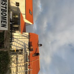 AS DAKWERKEN - Praca na dachu z czerwonej dachówki, widoczne rusztowania i dwóch dekarzy. Częściowo widoczny budynek z oknami i tablica z napisem 'KERSTBOMEN'. Pochmurne niebo.