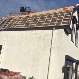 AS DAKWERKEN - Dach w trakcie krycia dachówką w Budel. Widoczna konstrukcja dachu z łatami i membraną. Na dole stosy dachówek. Prace dekarskie w toku, jasne niebo w tle.