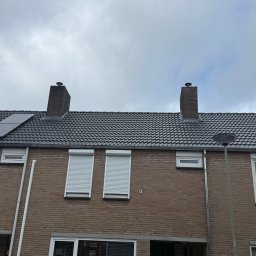 AS DAKWERKEN - Szary dach z dachówki ceramicznej na dwukondygnacyjnym domu z cegły, z dwoma kominami i roletami w oknach. Widok z dołu na pochmurne niebo.