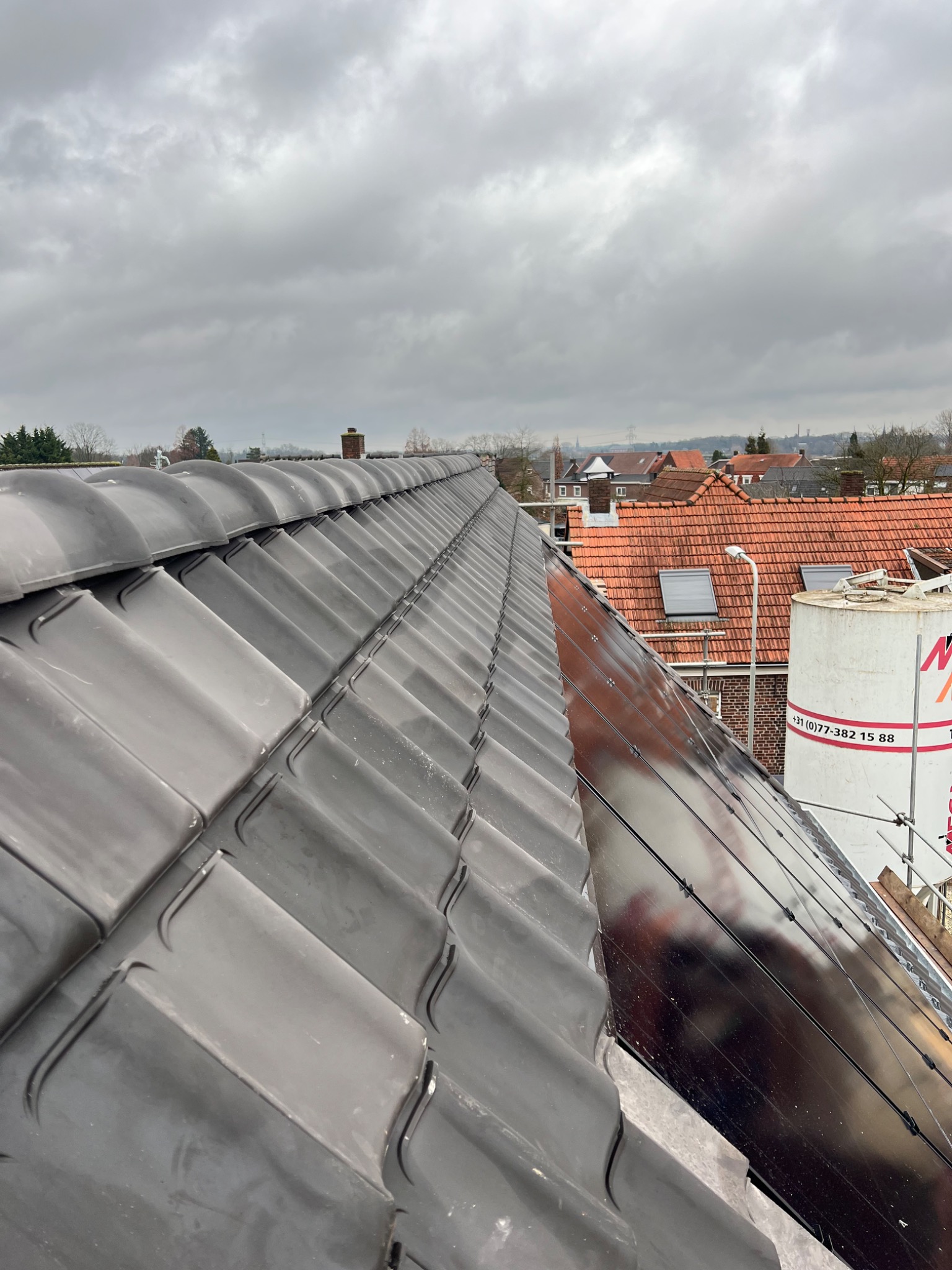 Widok z góry na fragment dachu pokrytego ciemną dachówką ceramiczną, z widocznymi panelami słonecznymi i fragmentem rusztowania w tle, w pochmurny dzień.