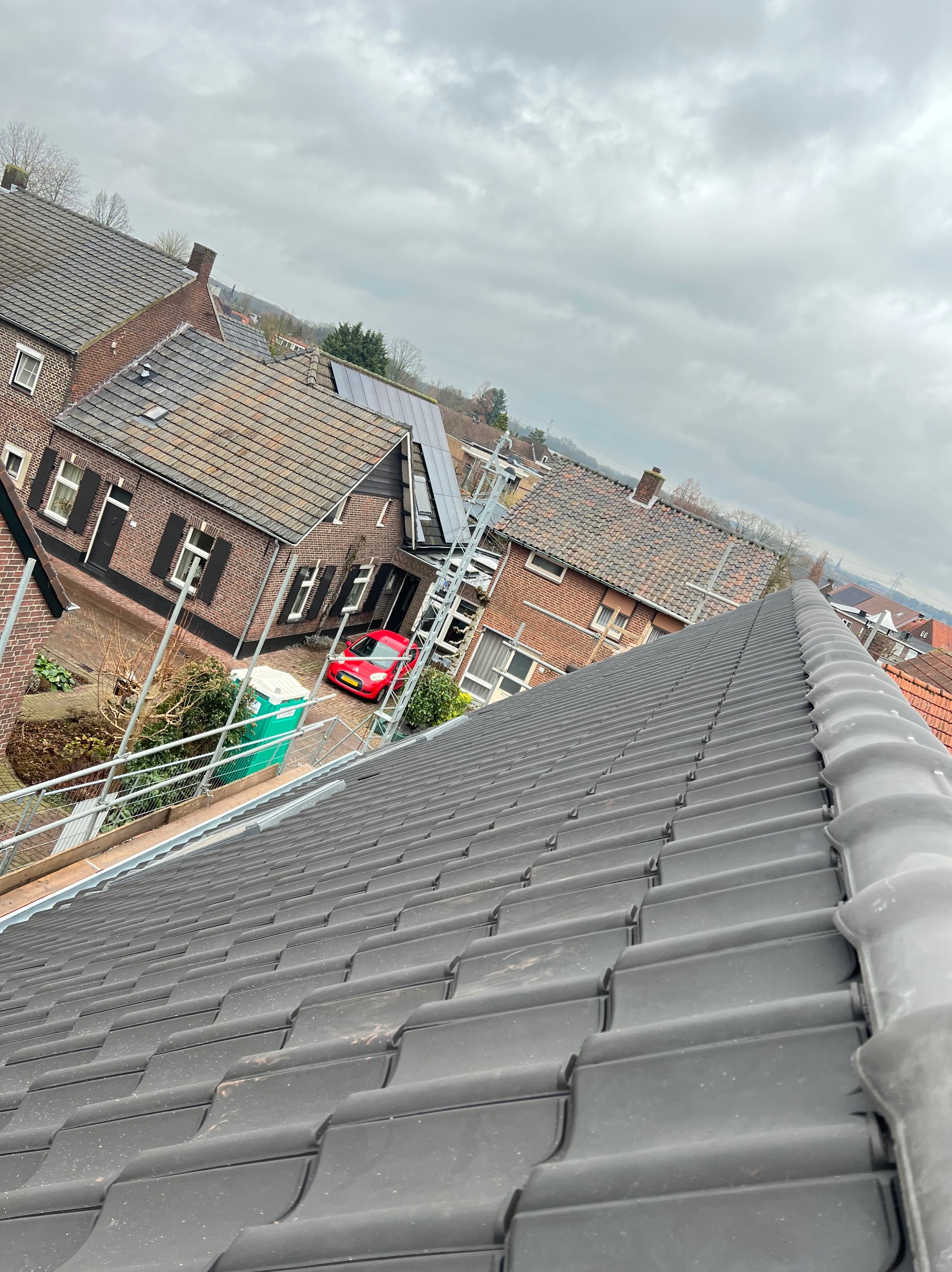 Nowy dach z ciemnoszarej dachówki ceramicznej, widok z góry na domy z czerwonej cegły i czerwoną osobówkę w tle. Metalowe rusztowanie. Pochmurny dzień.