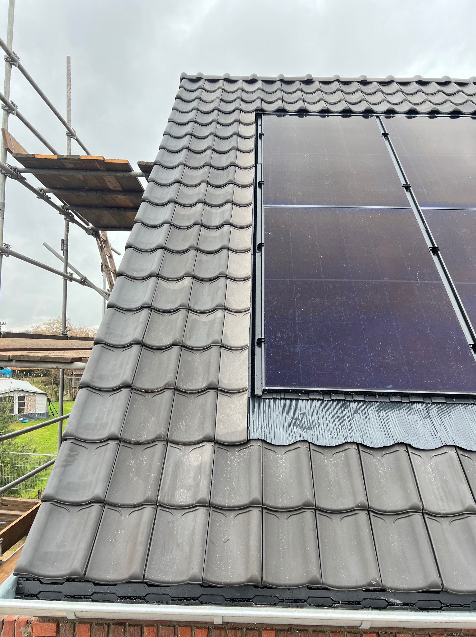 Montaż paneli solarnych na dachu z szarej dachówki ceramicznej. Widoczne rusztowanie i fragment ogrodu w tle. Dach po deszczu.