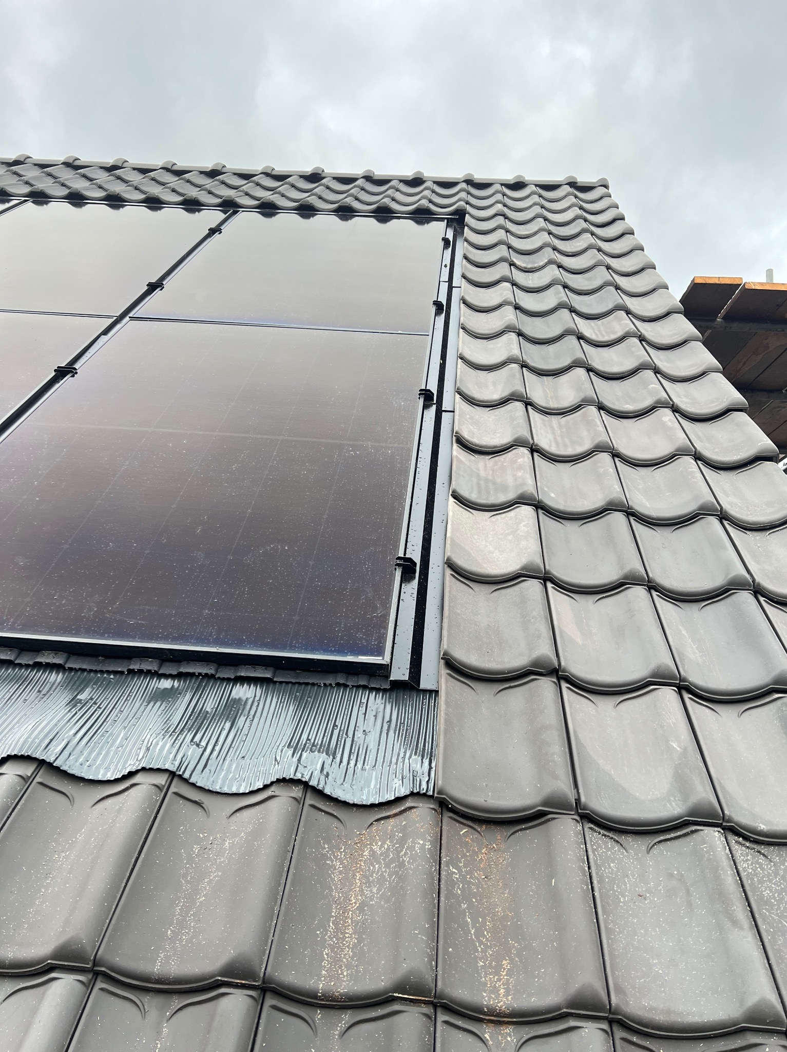 Montaż paneli słonecznych na dachu pokrytym szarą dachówką karpiówką. Widoczny fragment rusztowania. Ujęcie z dołu, pochmurne niebo.