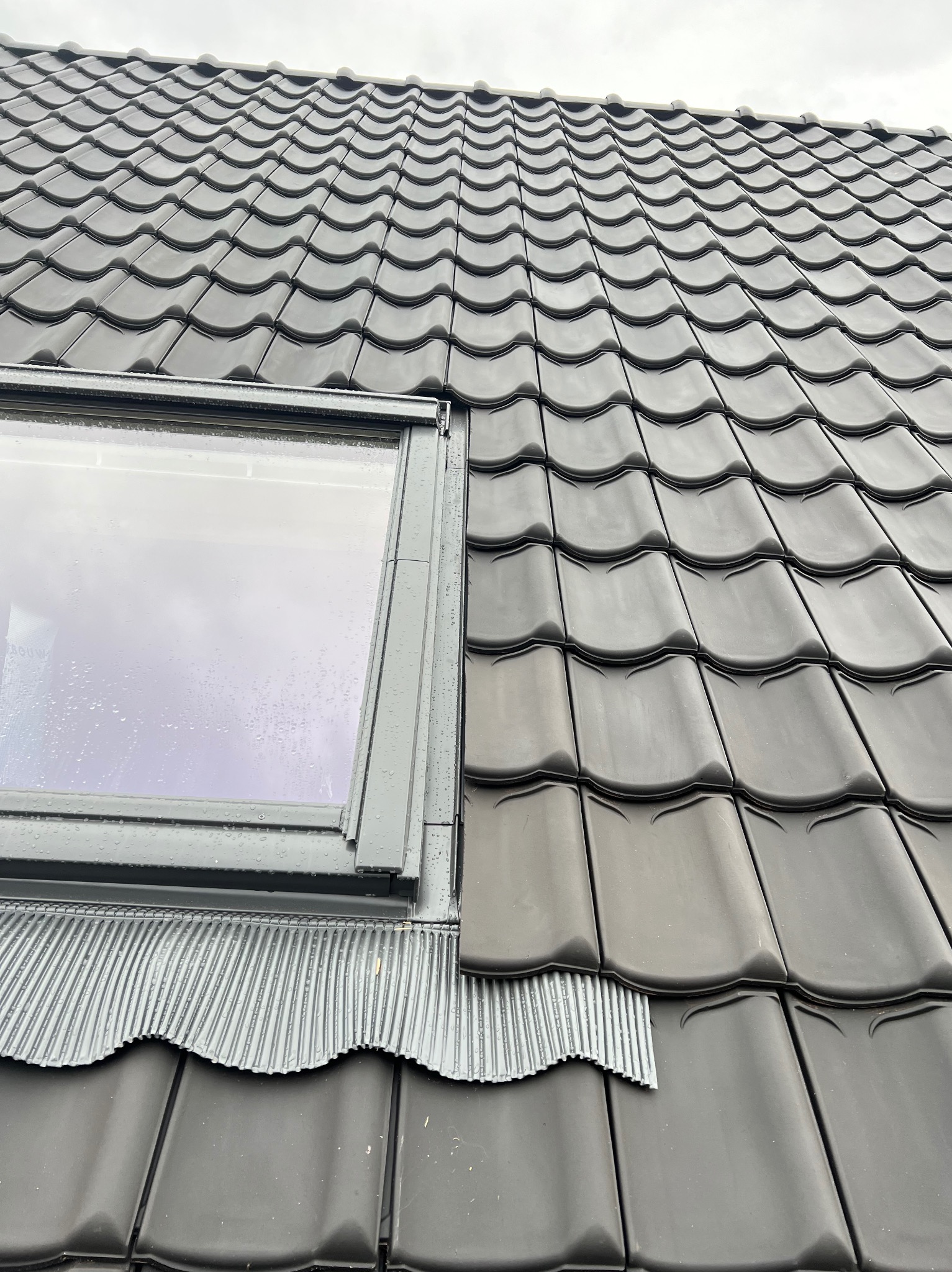 Szczegółowe ujęcie czarnej dachówki ceramicznej z oknem dachowym. Widoczne detale montażu oraz falisty kształt dachówki. Pochmurne niebo w tle.