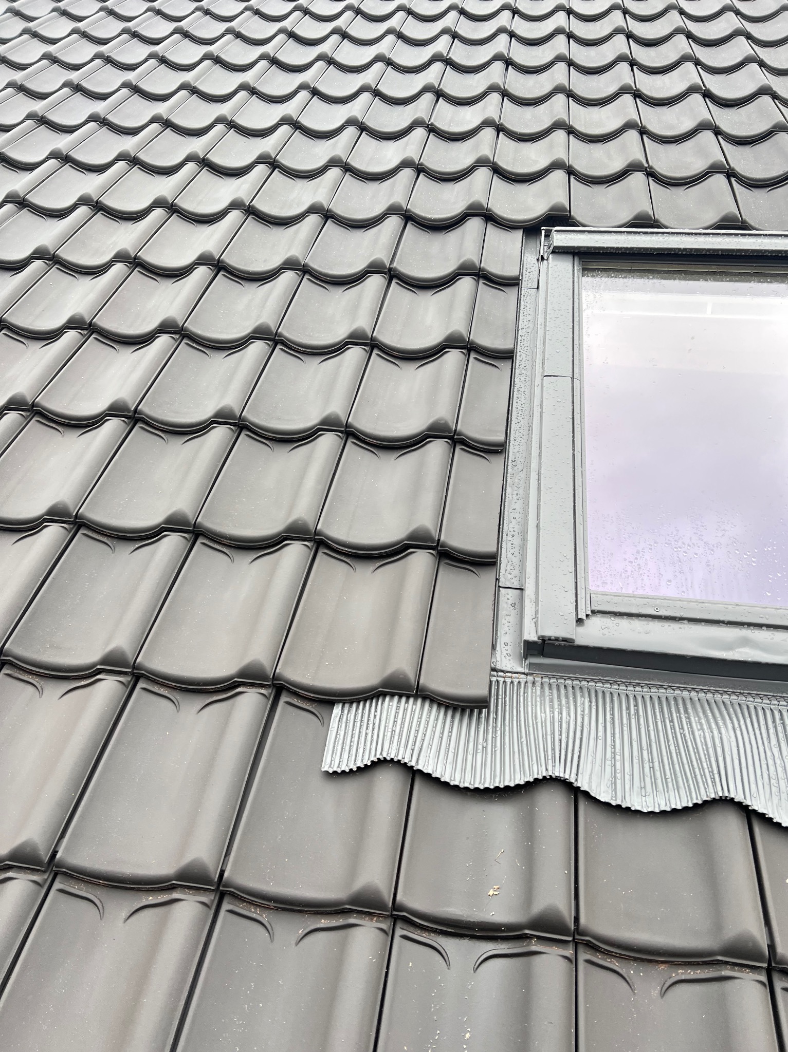 Nowe, grafitowe dachówki ceramiczne ułożone wokół okna dachowego z metalowym kołnierzem. Widok z góry, lekki deszcz.