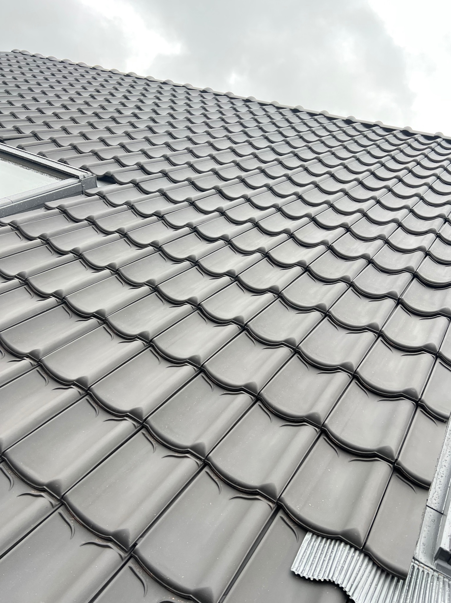 Szary dach z dachówki ceramicznej, widok z góry na połaci dachu z oknem dachowym i elementami wykończeniowymi. Pochmurne niebo w tle.