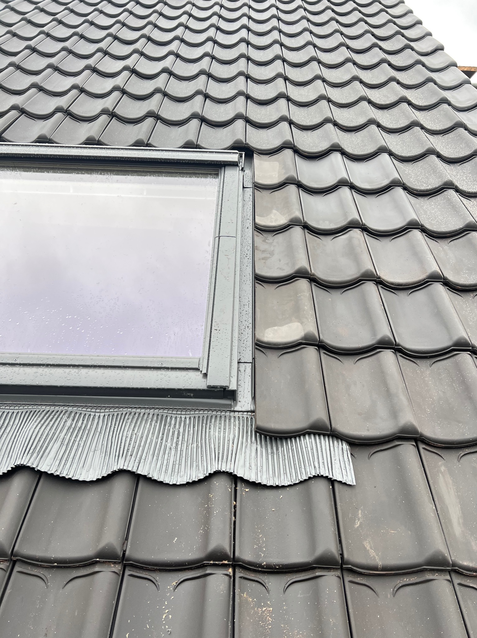 Czarna dachówka ceramiczna z oknem dachowym, widoczne elementy uszczelniające. Lekki deszcz na powierzchni, kadr z góry.