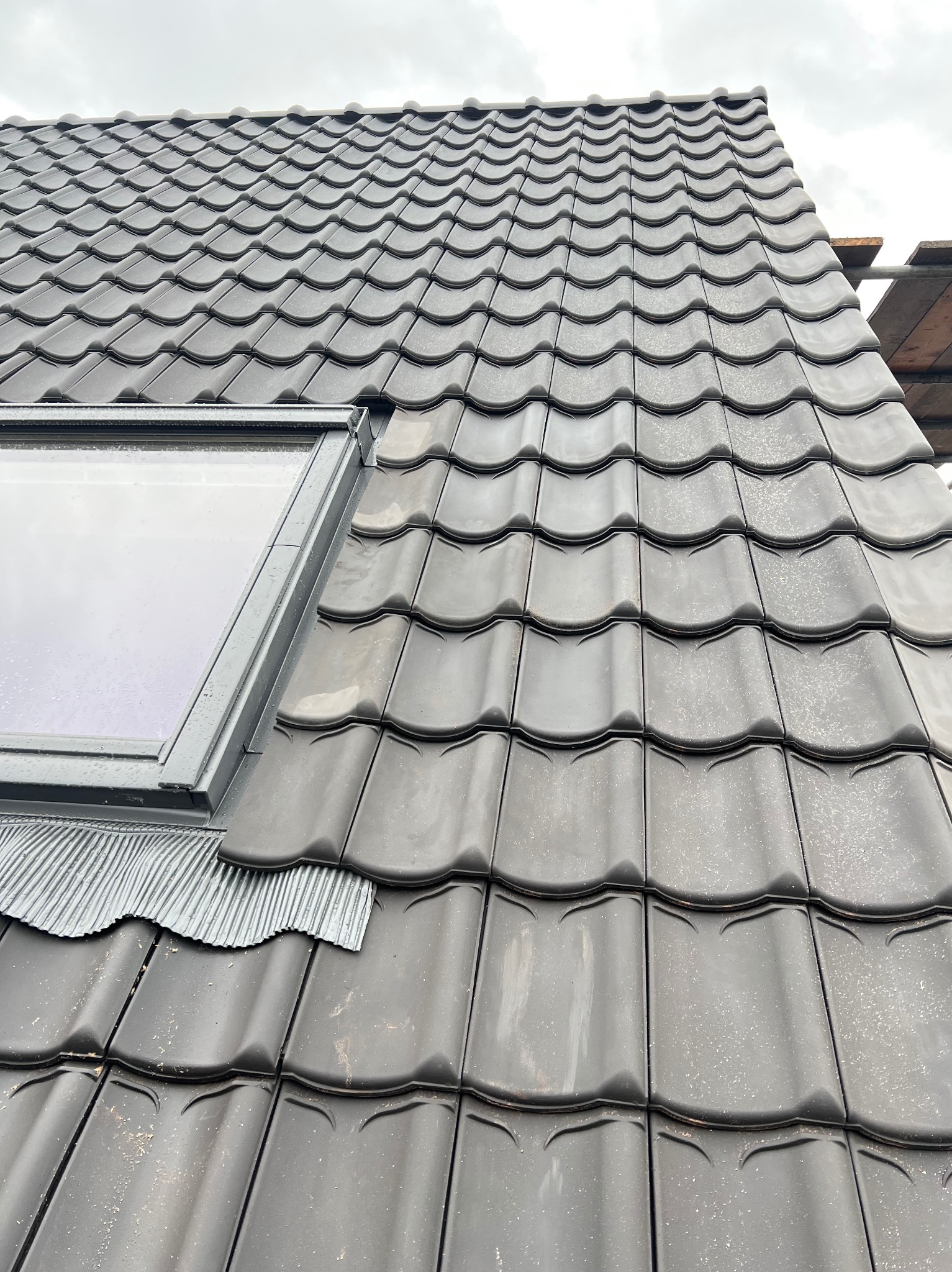 Szczegółowe ujęcie dachu z czarnej dachówki ceramicznej z oknem dachowym, widoczne elementy uszczelniające i fragment rusztowania. Pochmurne niebo w tle.