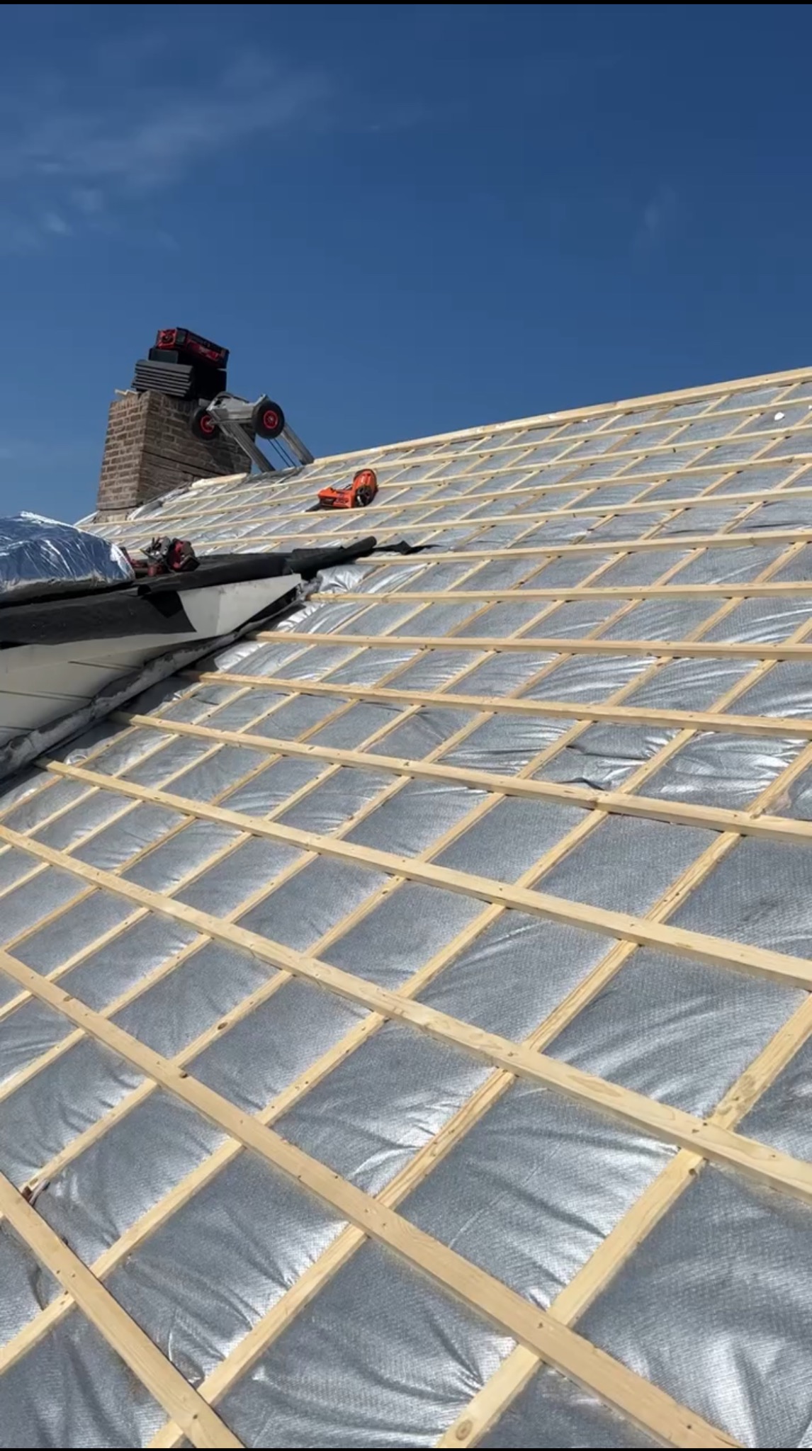 Konstrukcja dachu z izolacją, drewniane łaty tworzą kratownicę. Na kominie narzędzia dekarskie. Błękitne niebo w tle. Widok na dach z perspektywy.