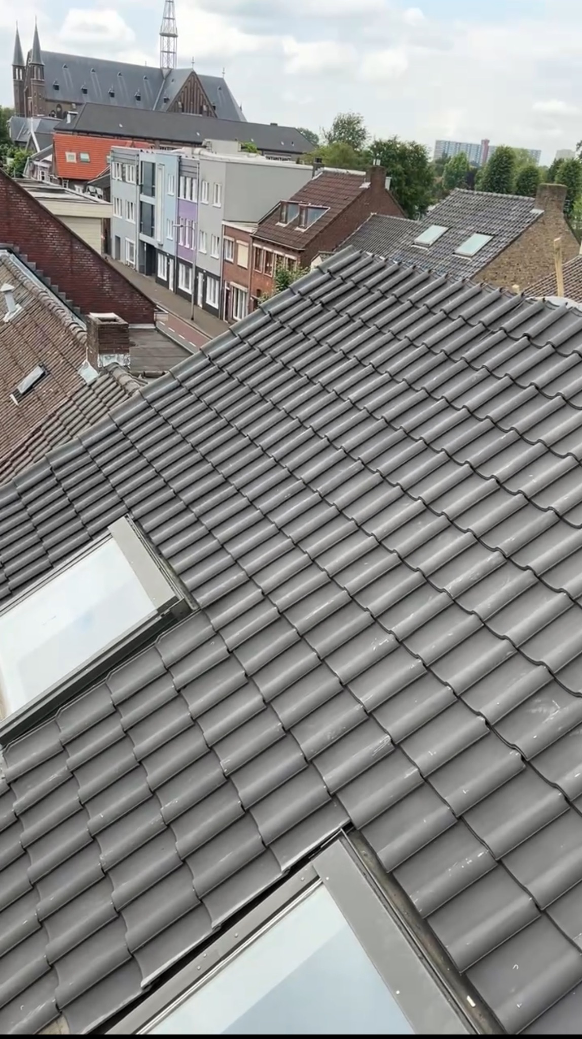 Szary dach z dachówkami i oknami dachowymi, widok z góry na tle zabudowy miejskiej i kościoła z wieżą.