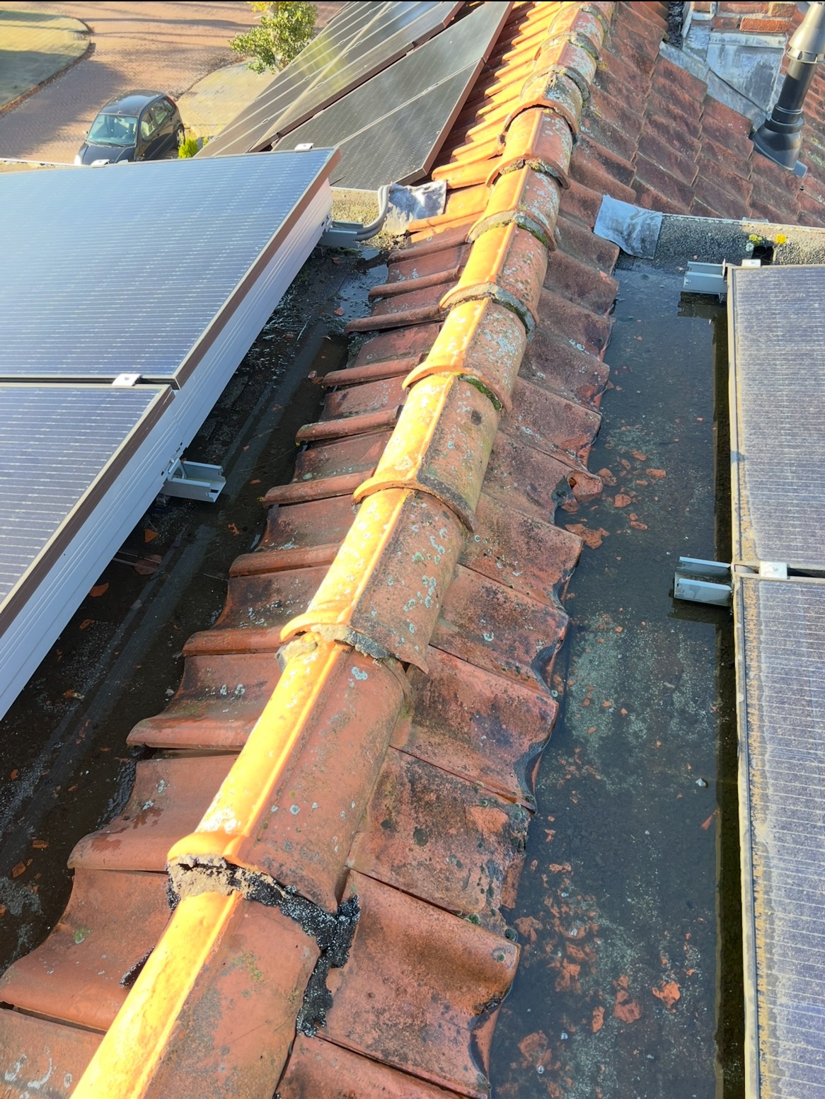 Czerwona dachówka karpiówka z mchem i wodą, obok panele słoneczne. Widoczne uszkodzenia i zabrudzenia na pokryciu dachowym. Ujęcie z góry.