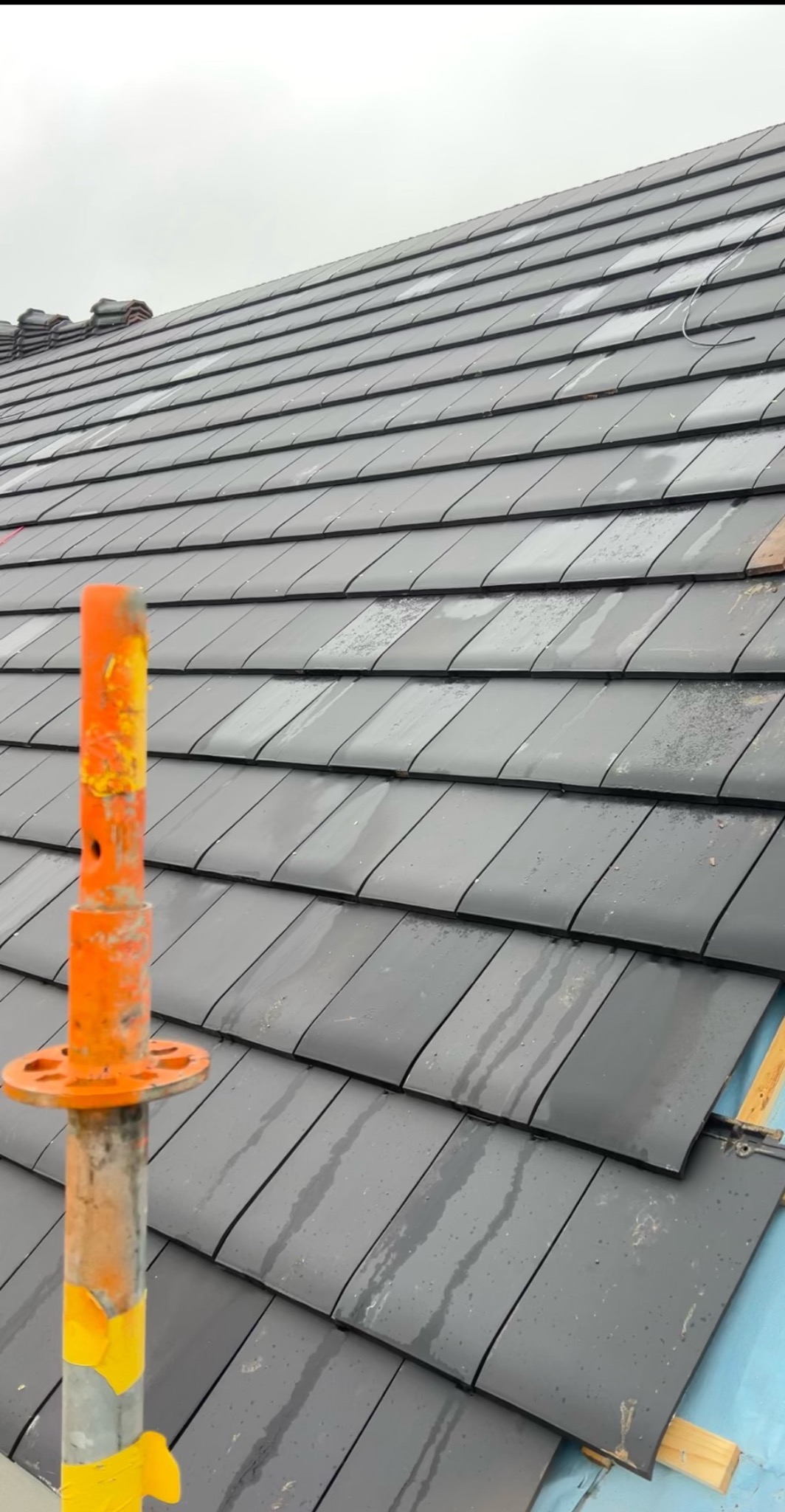 Ułożone, grafitowe dachówki na skośnym dachu, widoczne krople wody i pomarańczowe podpory. Dach pokryty folią, przygotowany do dalszych prac.