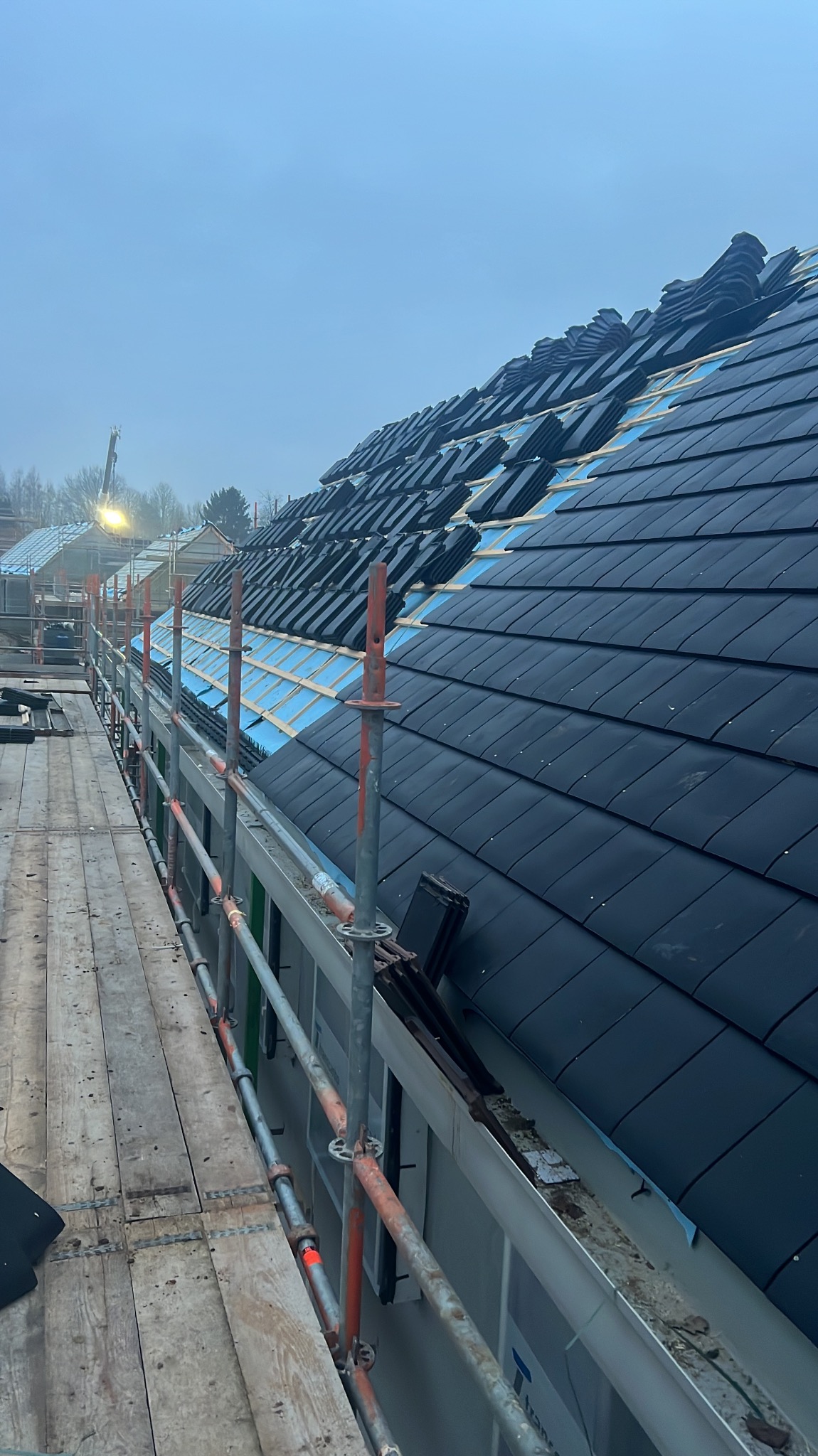 Częściowo pokryty dach czarną dachówką, widoczny z rusztowania. Niebieska membrana dachowa i ułożone rzędy dachówek czekają na dokończenie krycia.