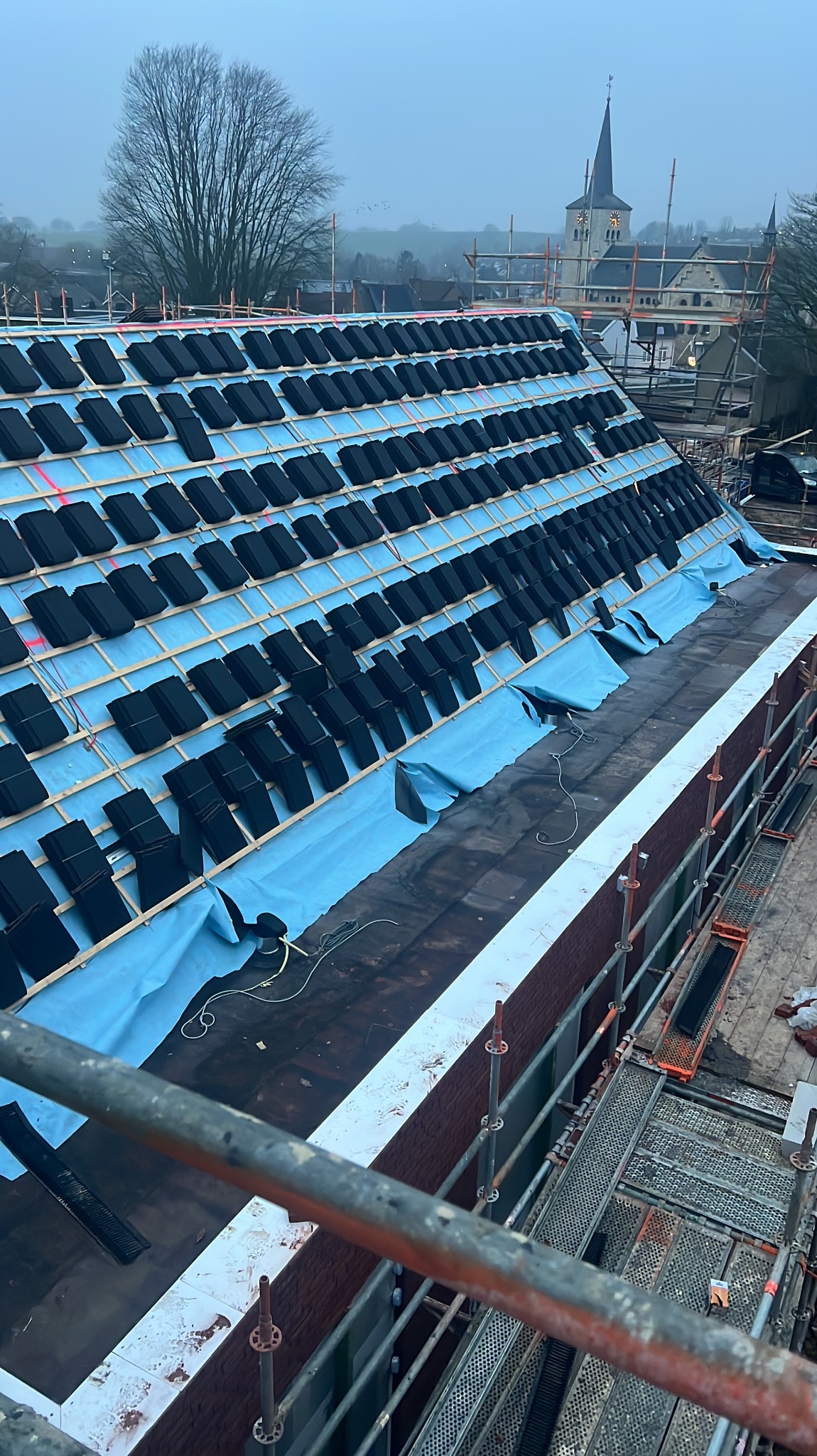 Układanie czarnej dachówki na zabezpieczonej folią membranie dachu. Widoczne drewniane łaty, rusztowania i oddalony kościół z wieżą w tle.