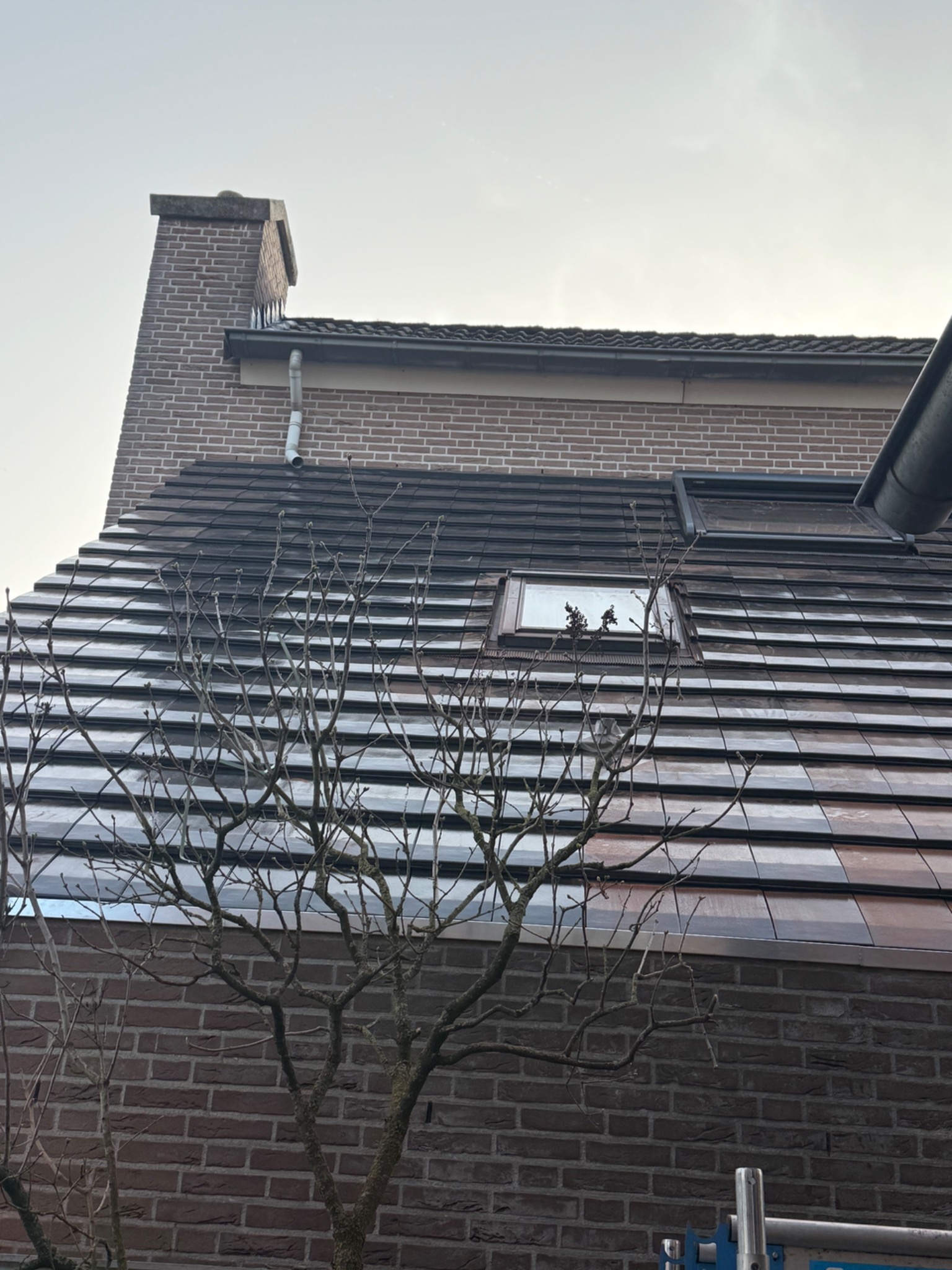 Dachówka ceramiczna w odcieniach brązu i szarości na skośnym dachu domu z cegły, widoczny komin i okno dachowe, gałęzie drzewa na pierwszym planie.