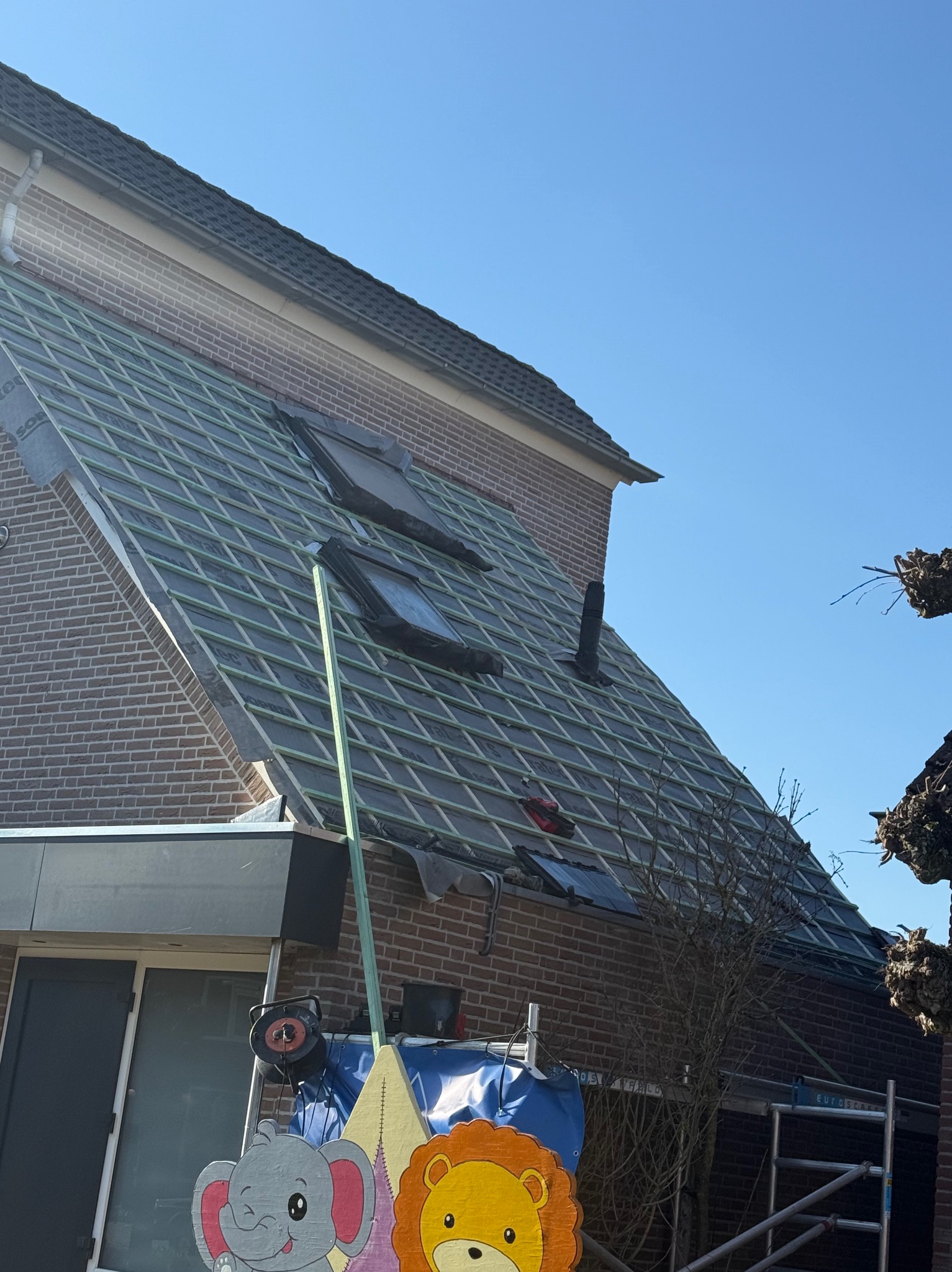 Dach z oknami połaciowymi w trakcie krycia, widoczne łaty i kontrłaty. Częściowo pokryty dachówką, rusztowanie z narzędziami i dekoracjami w tle.