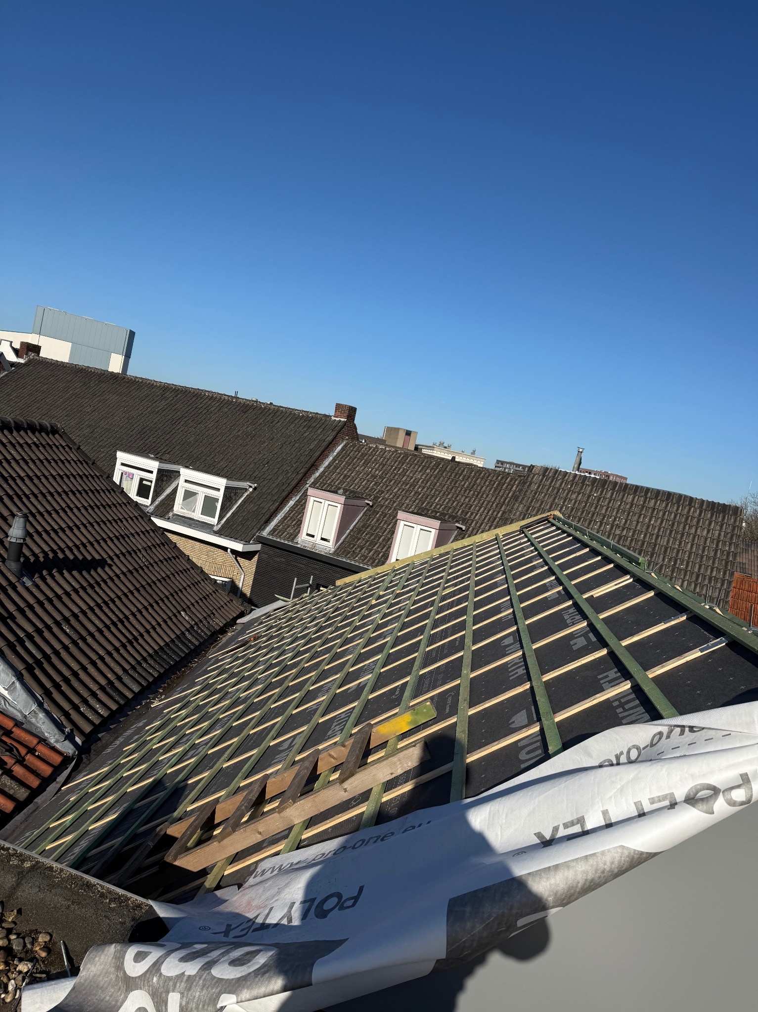 Konstrukcja dachu z łatami i membraną w trakcie montażu, widok z góry na tle innych dachów i błękitnego nieba. Częściowo widoczna folia ochronna.