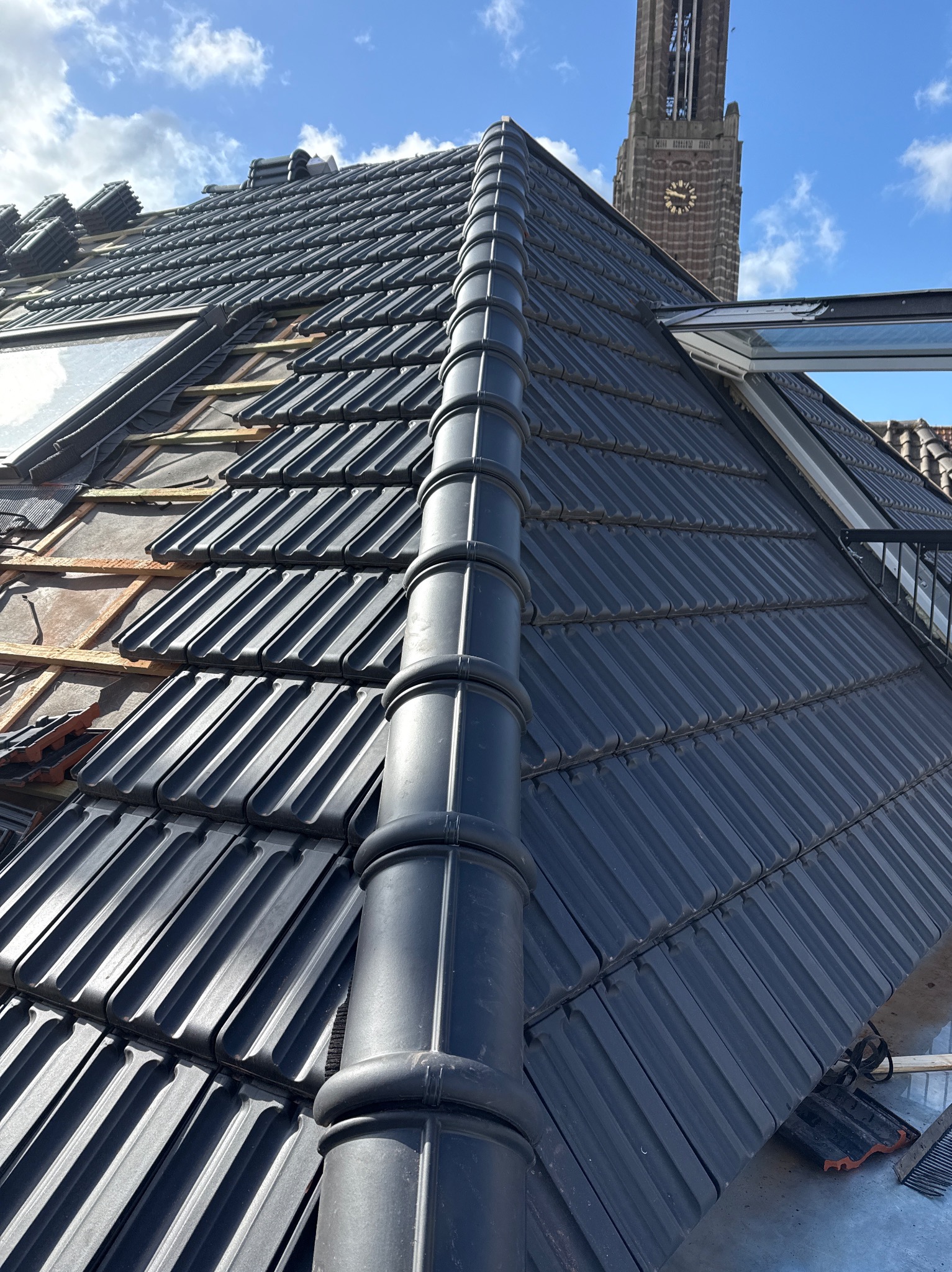 Częściowo pokryty dach z czarnej dachówki ceramicznej, widoczne okna dachowe i fragment wieży zegarowej w tle. Nowe pokrycie dachu w trakcie realizacji.