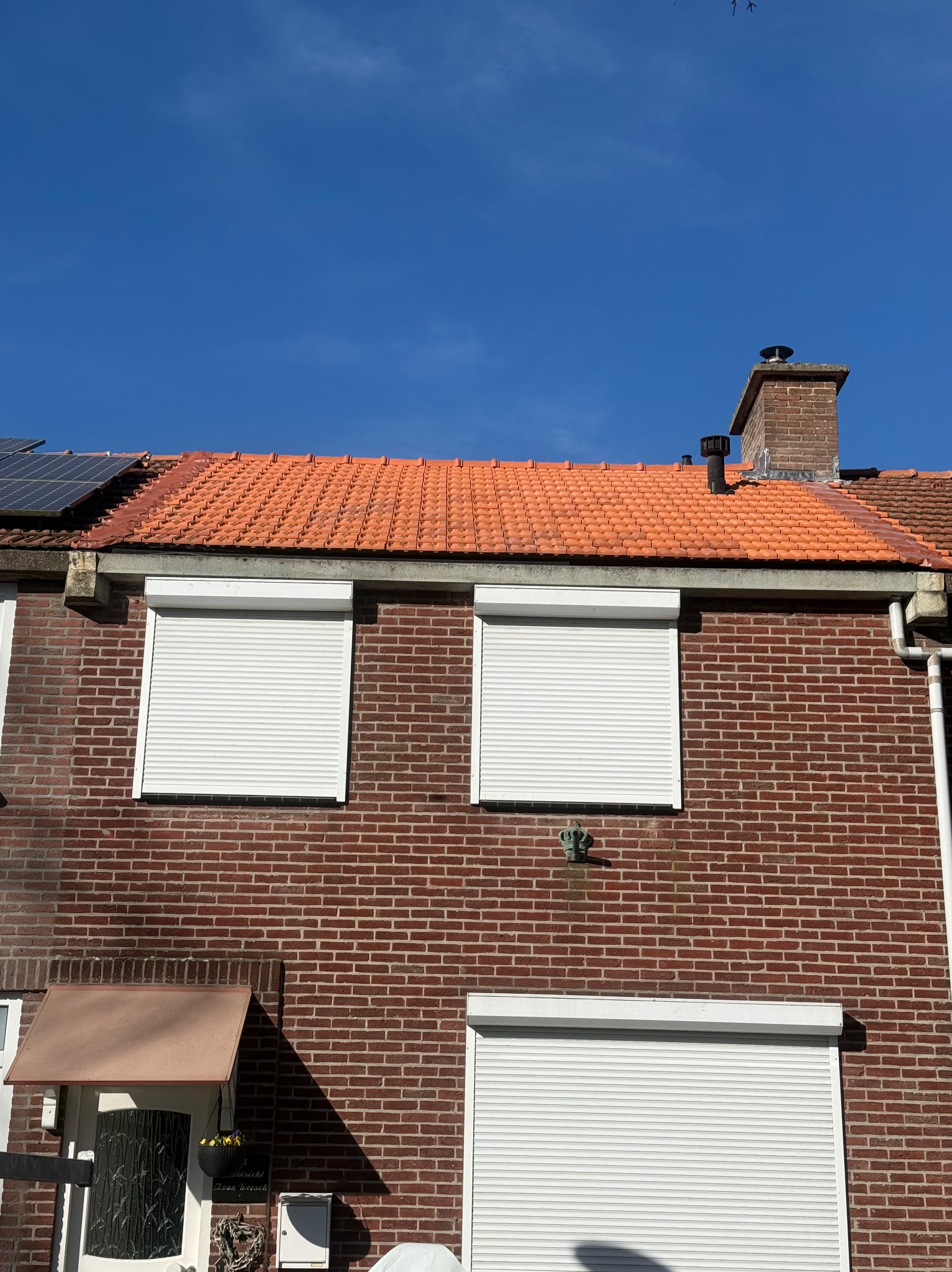 Ceglany dom z pomarańczową dachówką i białymi roletami okiennymi w słoneczny dzień. Widoczny komin i fragment paneli słonecznych na dachu.