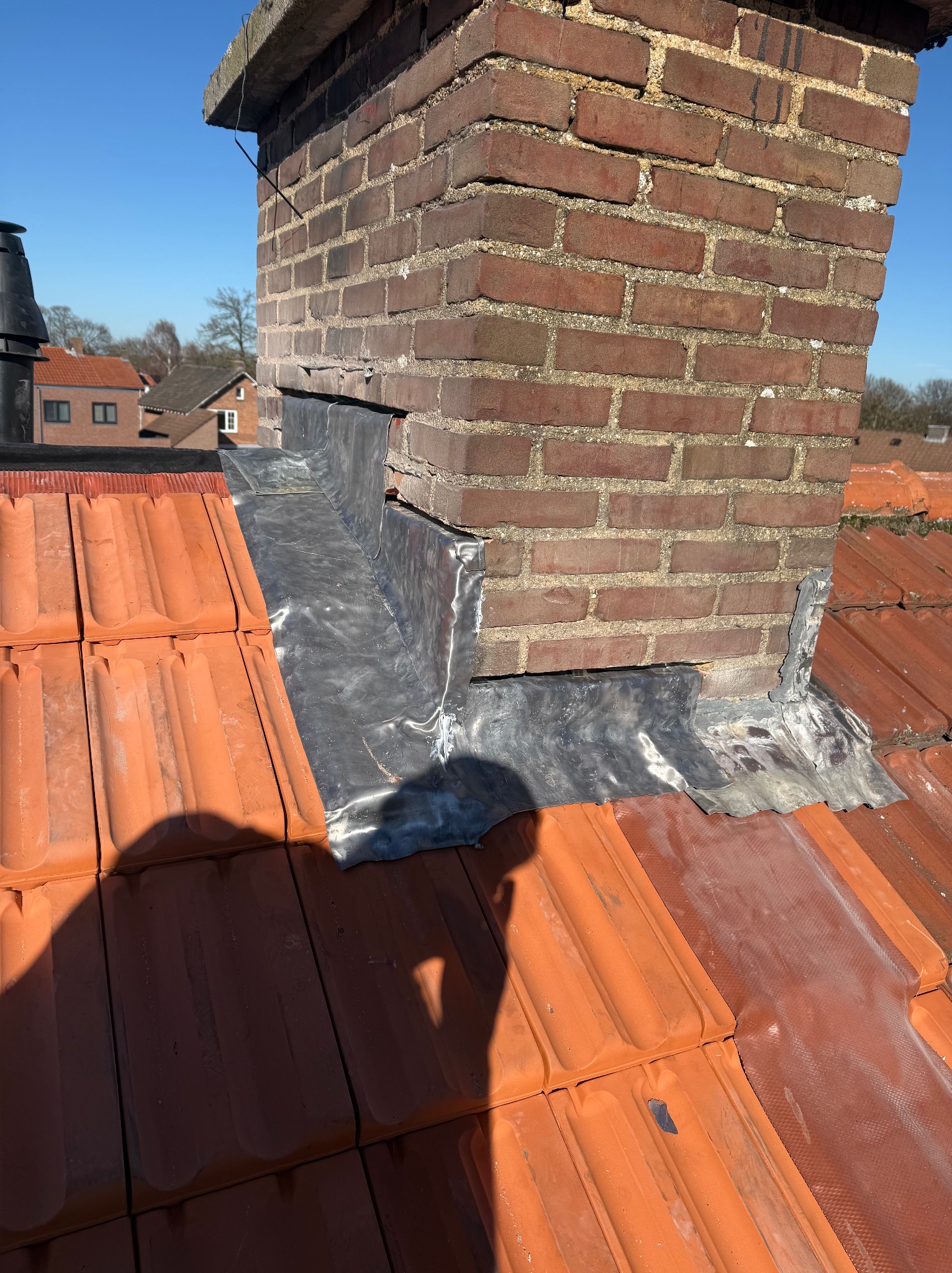 Ceglany komin z obróbką blacharską na dachu z czerwonej dachówki, widok z góry w słoneczny dzień. Detal zabezpieczenia komina przed przeciekaniem.