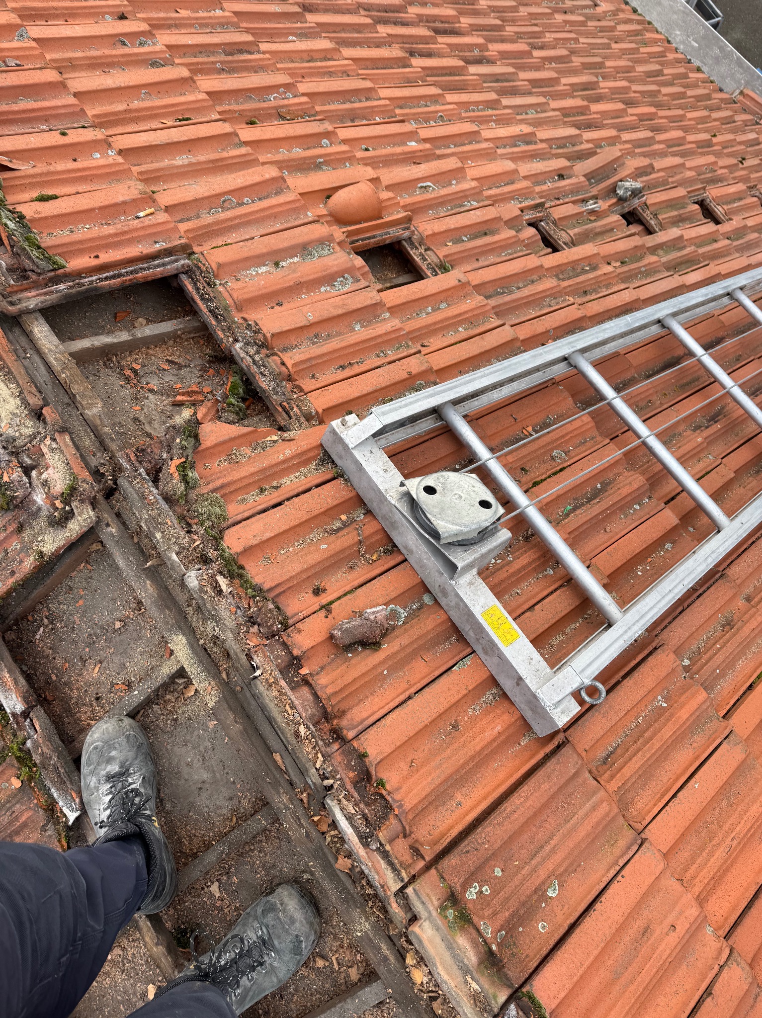 Częściowo zdjęty, stary dach z czerwonej dachówki ceramicznej. Widoczne uszkodzenia, mech i drabina dekarska. Ujęcie z góry, z perspektywy osoby stojącej na dachu.