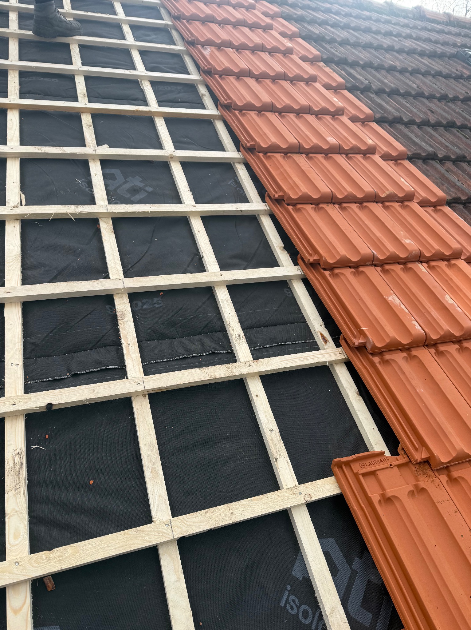 Częściowo pokryty dach czerwoną dachówką, widoczne łaty i membrana dachowa. Prace dekarskie w Budel. Dachówka Laumans.