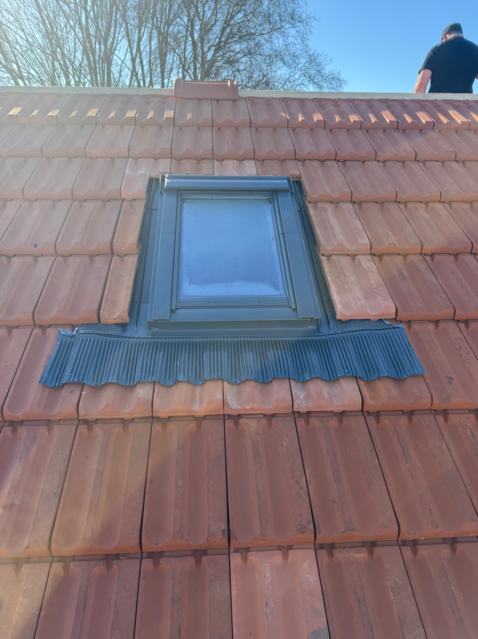 Czerwona dachówka ceramiczna z oknem dachowym i specjalnym kołnierzem uszczelniającym. Widok z góry, fragment dachu i sylwetka dekarza w tle.