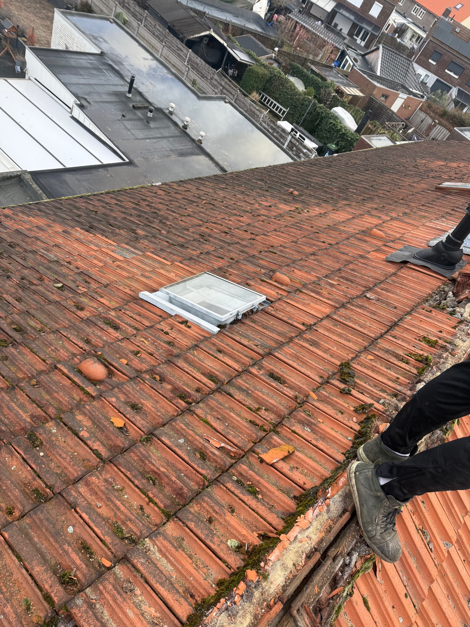 Czerwona dachówka z mchem i fragmentem okna dachowego. Widoczne buty dekarza na krawędzi dachu. Ujęcie z góry, ukośne.