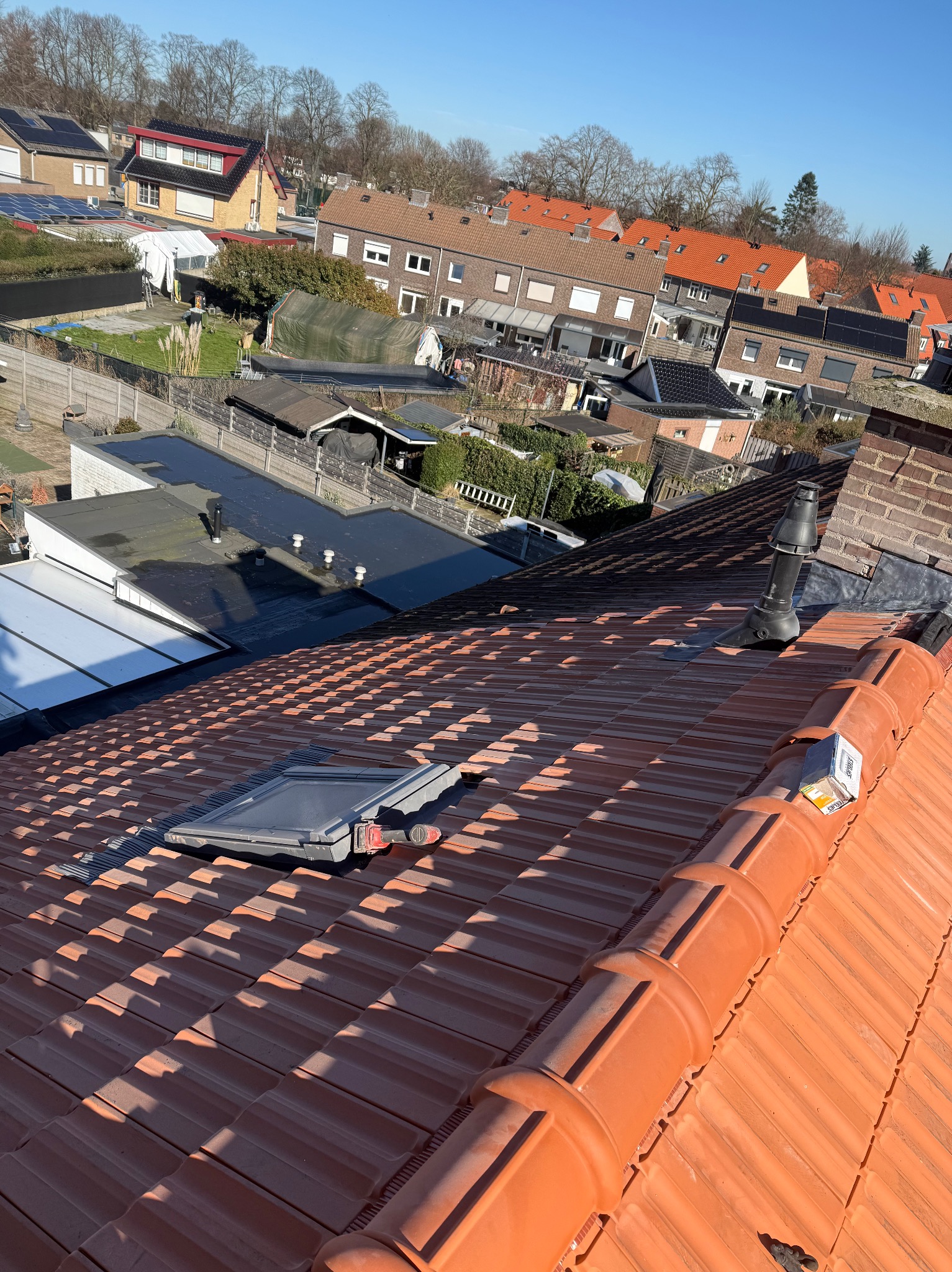 Czerwona dachówka ceramiczna z oknem dachowym i kominkiem wentylacyjnym na tle zabudowy jednorodzinnej w słoneczny dzień. Widok z góry.