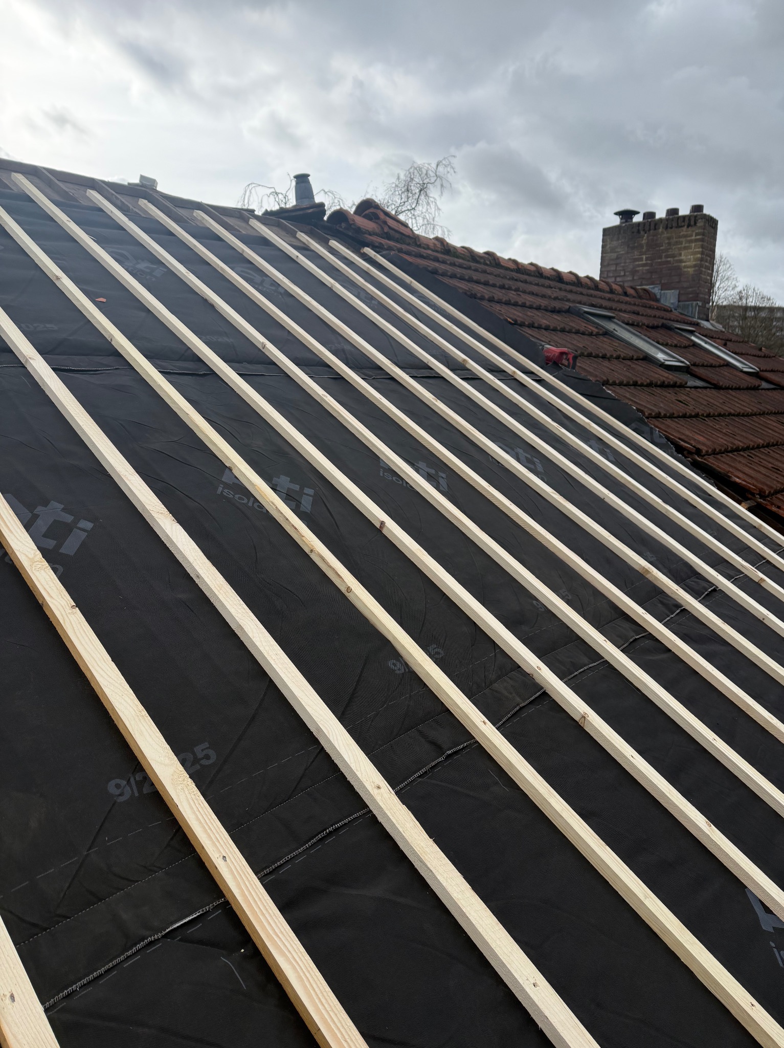 Częściowy widok dachu z łatami i membraną izolacyjną, przygotowanego pod nowe pokrycie. W tle komin i fragment starej dachówki.