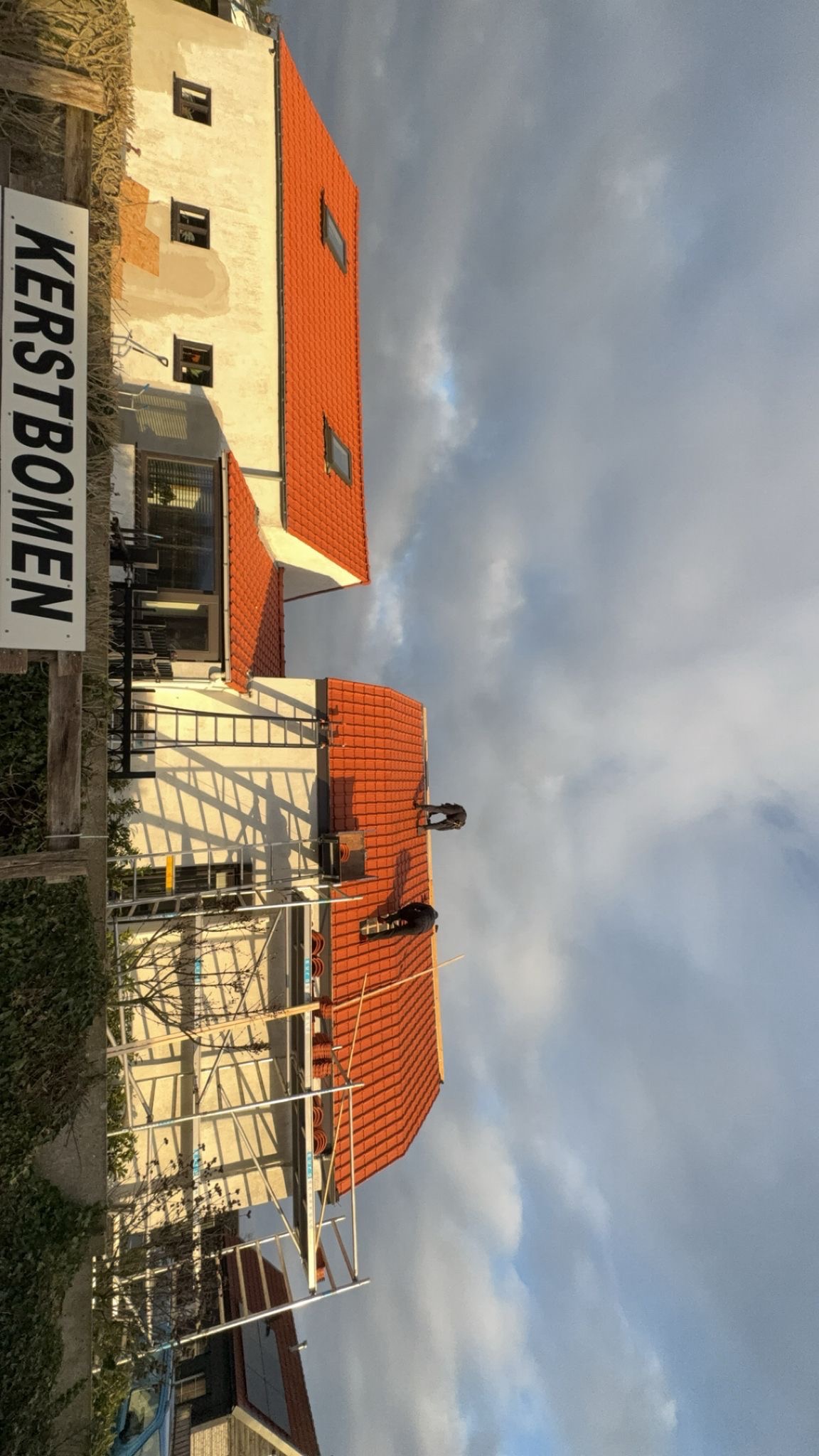 Praca na dachu z czerwonej dachówki, widoczne rusztowania i dwóch dekarzy. Częściowo widoczny budynek z oknami i tablica z napisem 'KERSTBOMEN'. Pochmurne niebo.